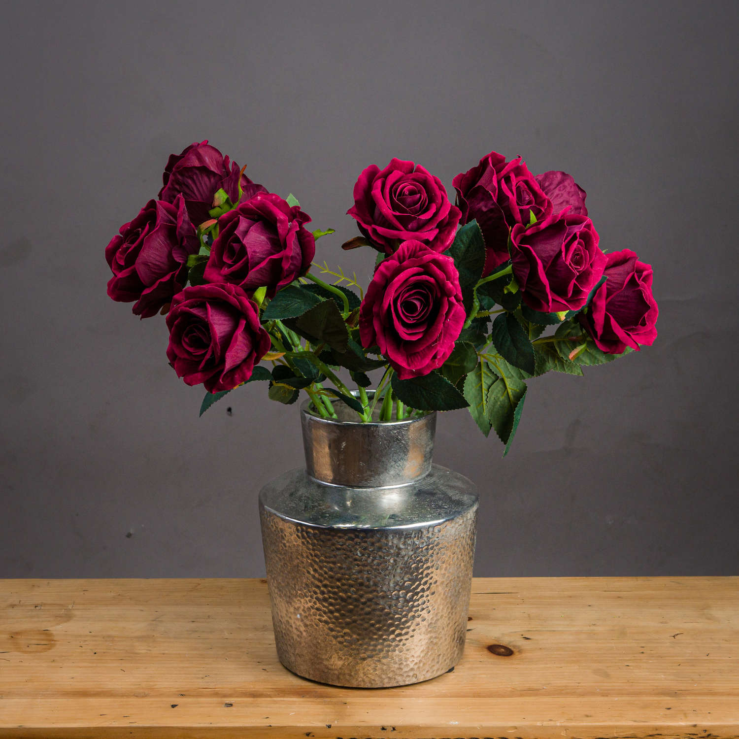 Deep red artificial roses in a hammered silver vase, showcasing velvety petals and lush green leaves, adding elegance to any decor.