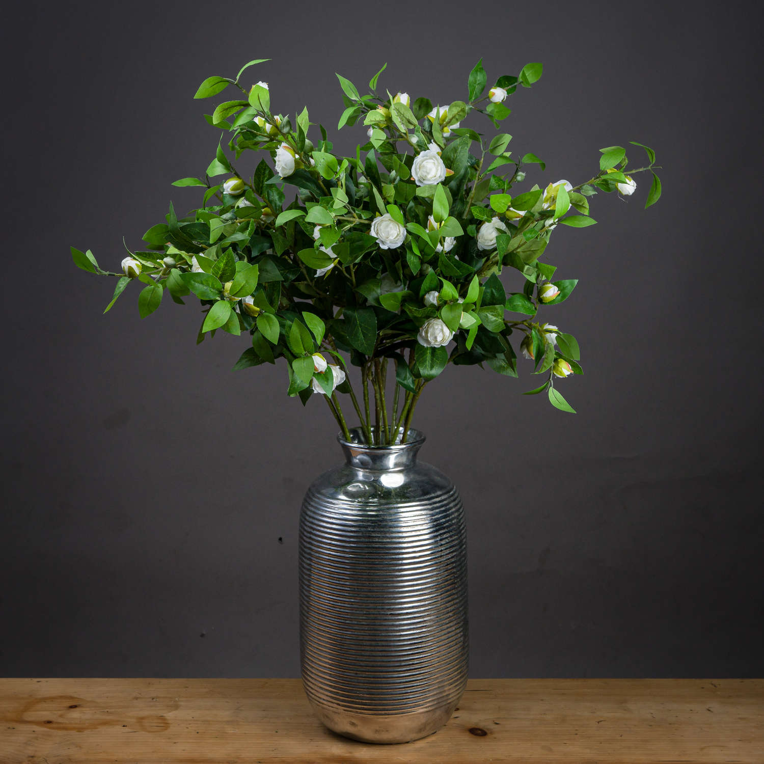 Cream gardenia stem with realistic cream petals and green leaves, arranged in a textured silver vase against a dark background.