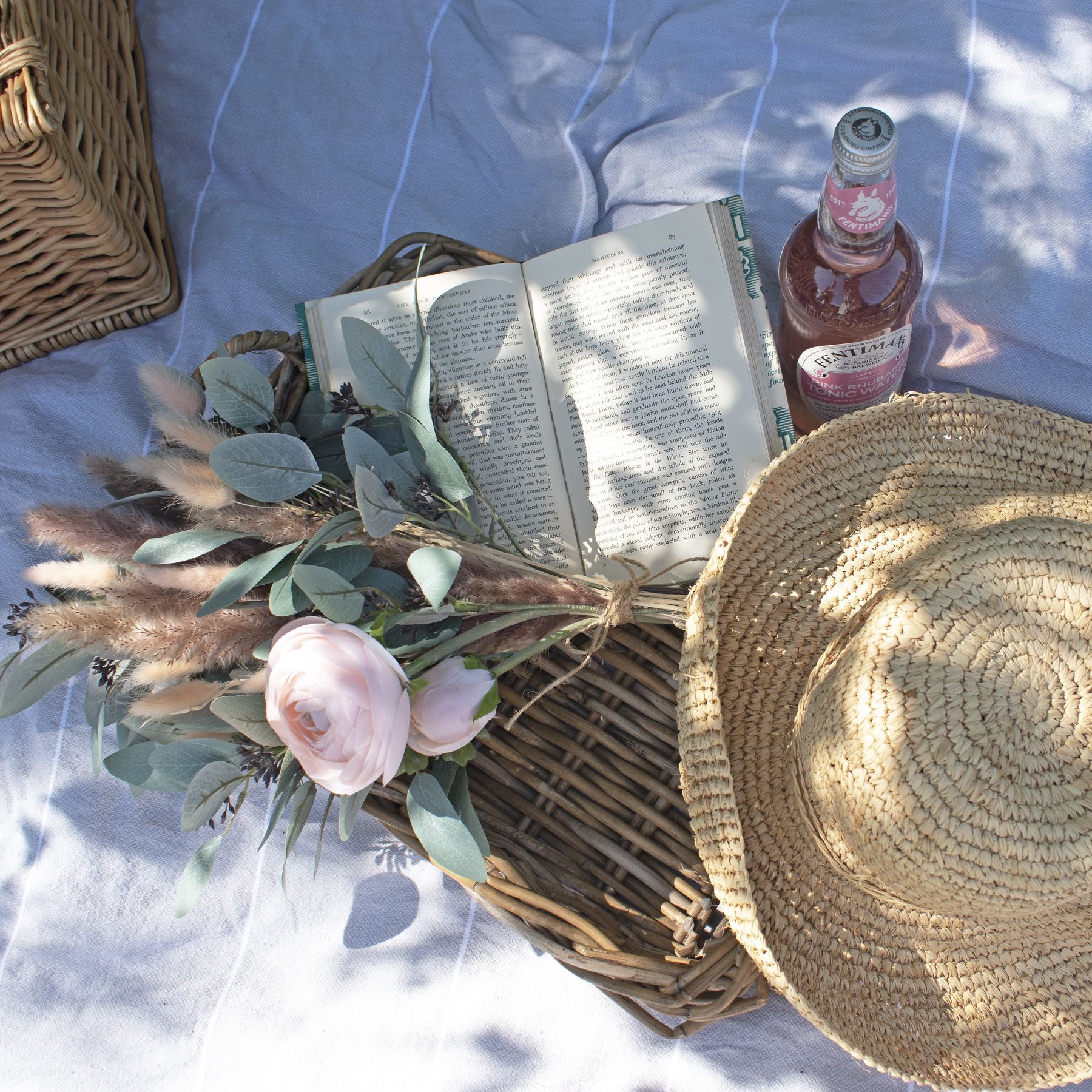Pastel pink ranunculus blooms with delicate petals, surrounded by green foliage, set on a textured surface with a book and straw hat.