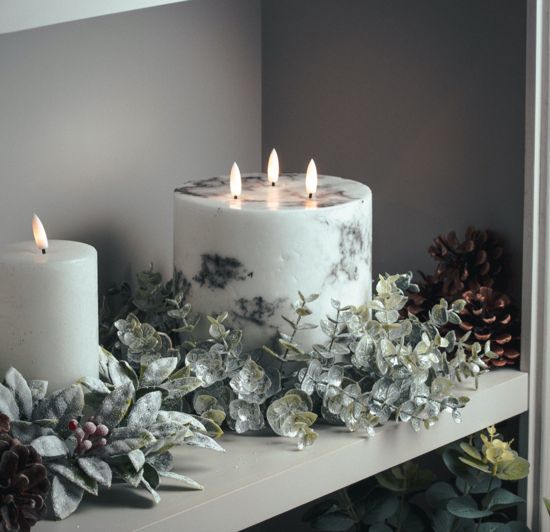 White LED candles with a marble effect glow warmly among greenery and pinecones, creating an elegant ambiance.