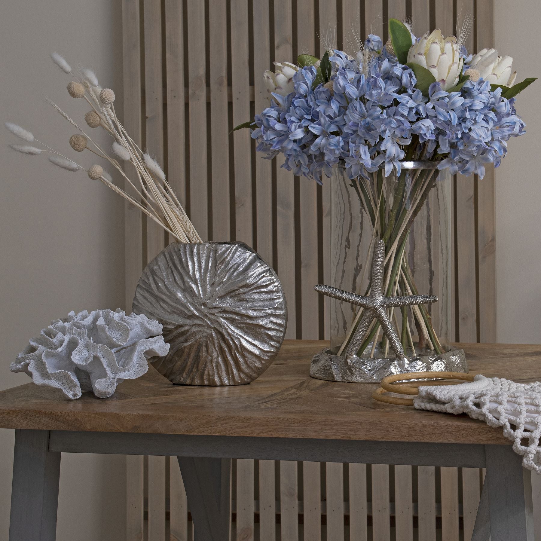 Light blue agapanthus flowers in a clear vase, complemented by silver decor and natural elements, creating a serene coastal ambiance.