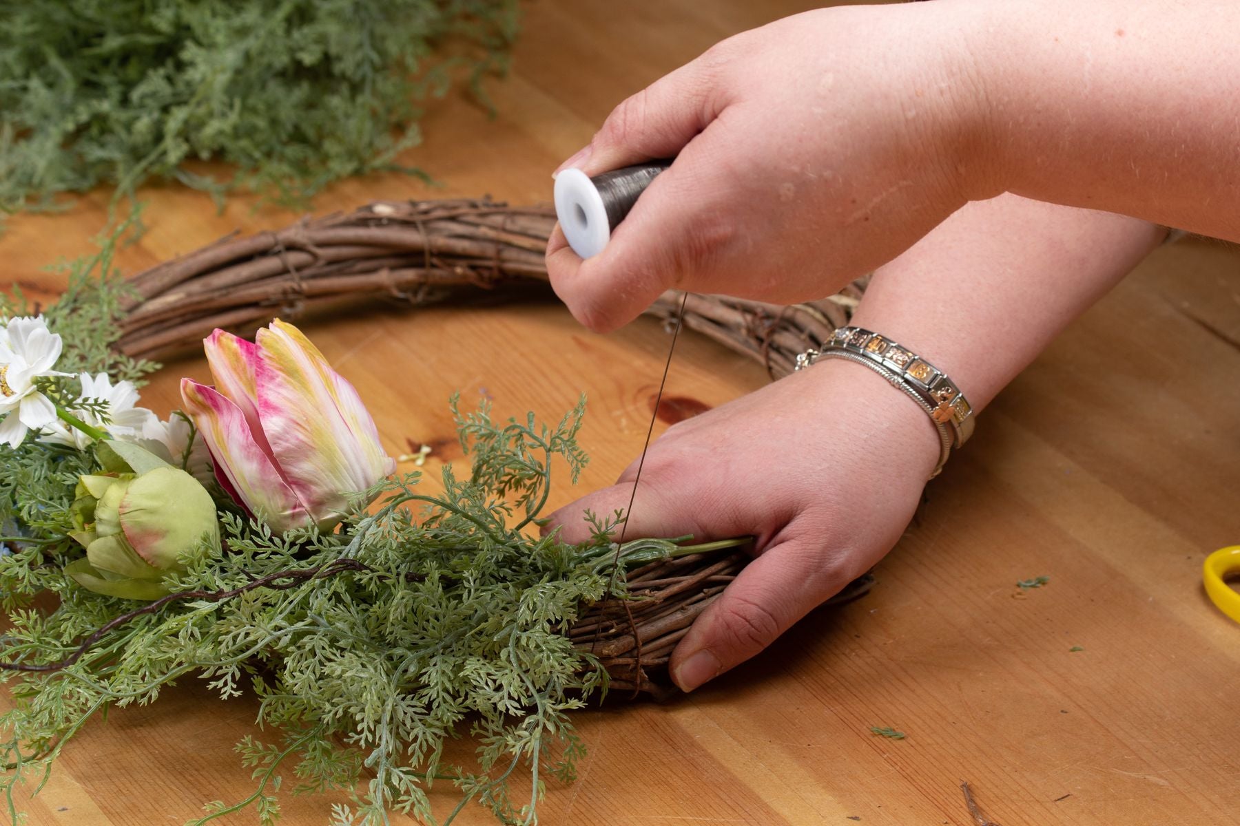 Hand arranging a vibrant Asparagus Fern bunch with feathery green fronds, alongside pink and white artificial flowers on a wooden table.