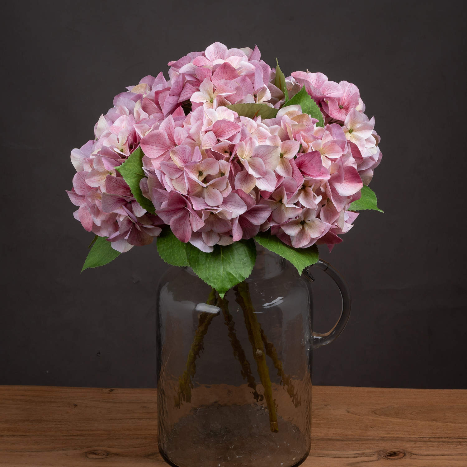 Shabby pink artificial hydrangea blooms with subtle color variations in a glass vase, accented by green leaves.