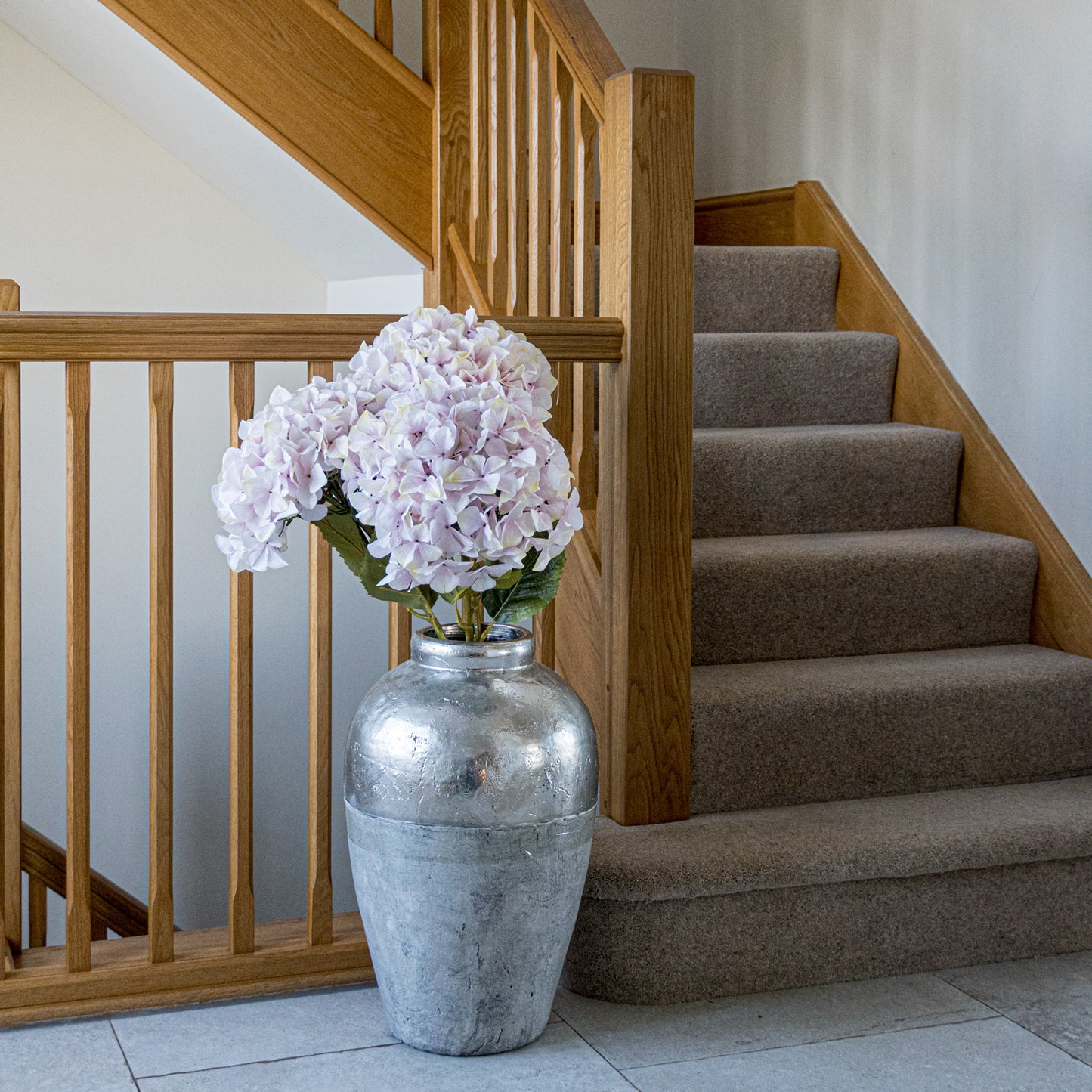 Full-bloom artificial pink hydrangea in a silver vase, elegantly displayed near a wooden staircase, enhancing interior decor.