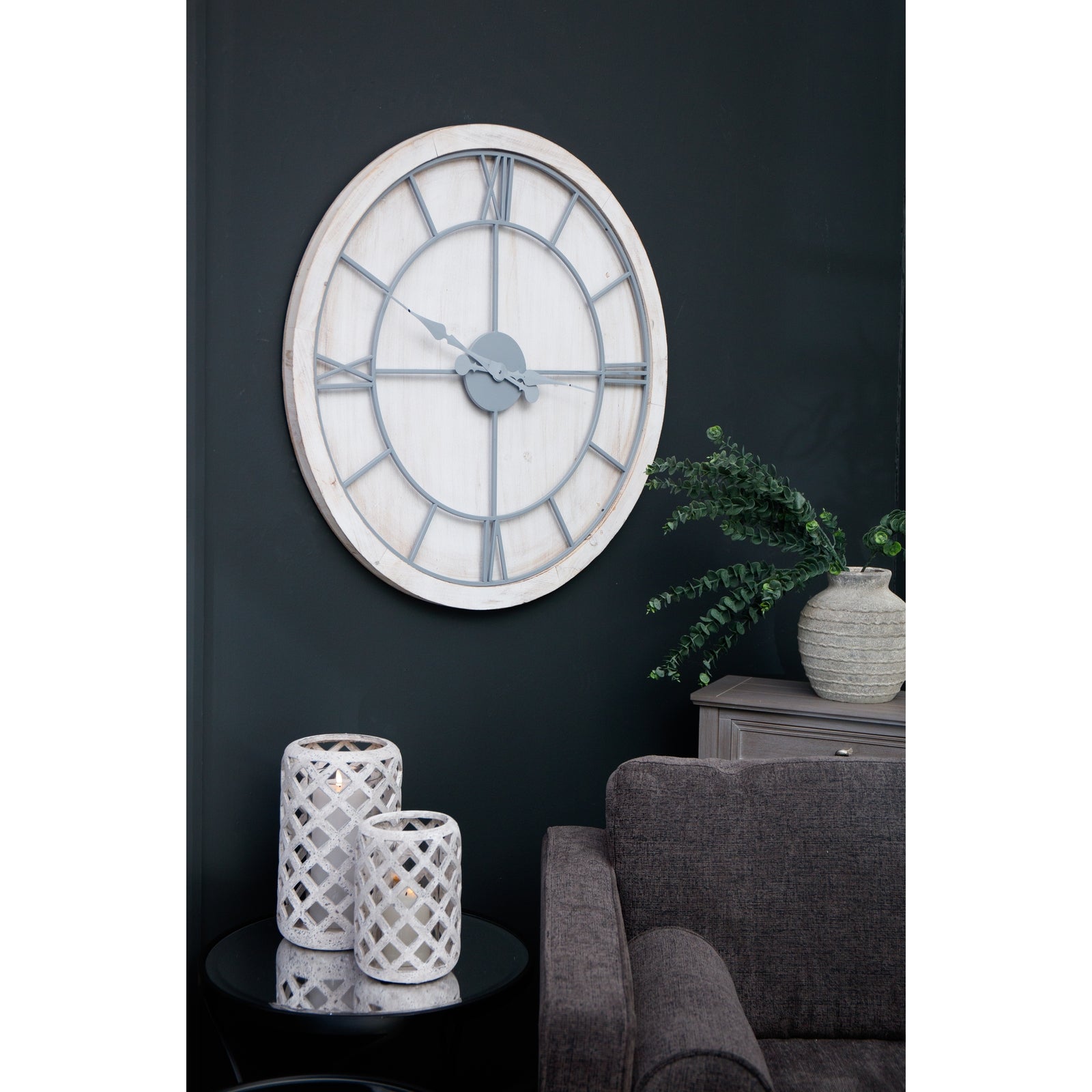 Large white wall clock featuring a round whitewashed wood frame and light grey Roman numerals, mounted on a dark wall.