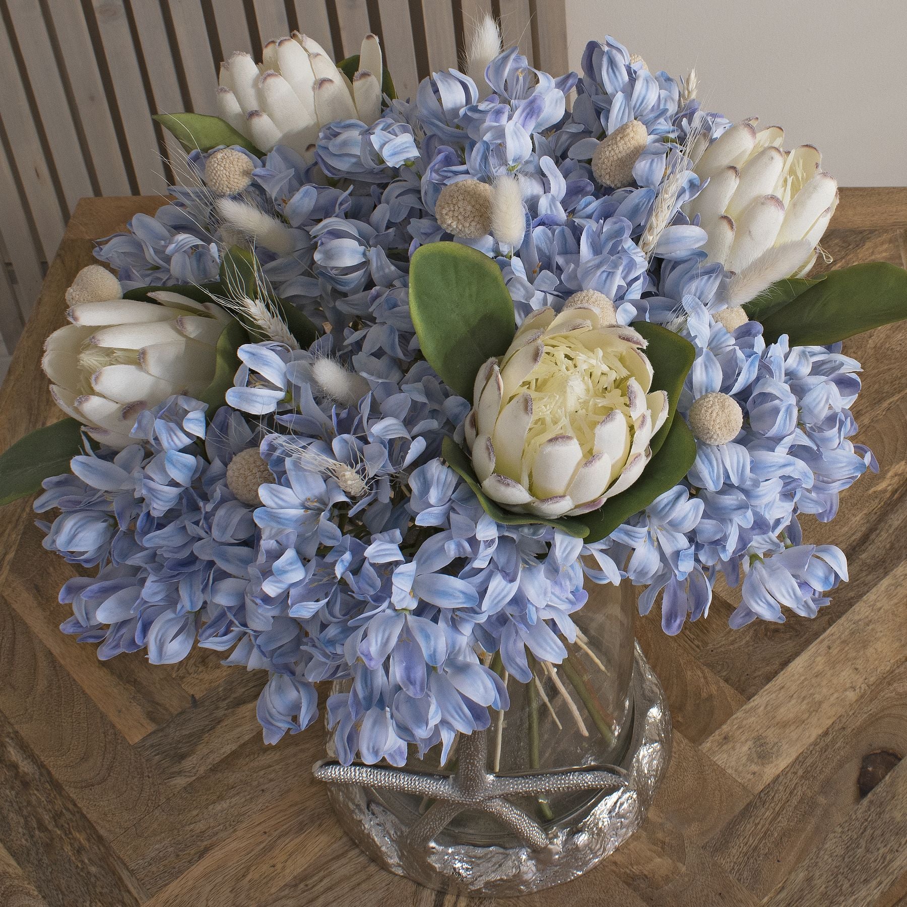 Bunch of dried white bunny tail stems featuring soft, delicate plumes, arranged with blue flowers in a glass vase.