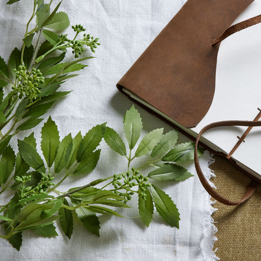 Long artificial fennel stem with intricate green foliage, resting beside a brown leather notebook on a textured white linen background.