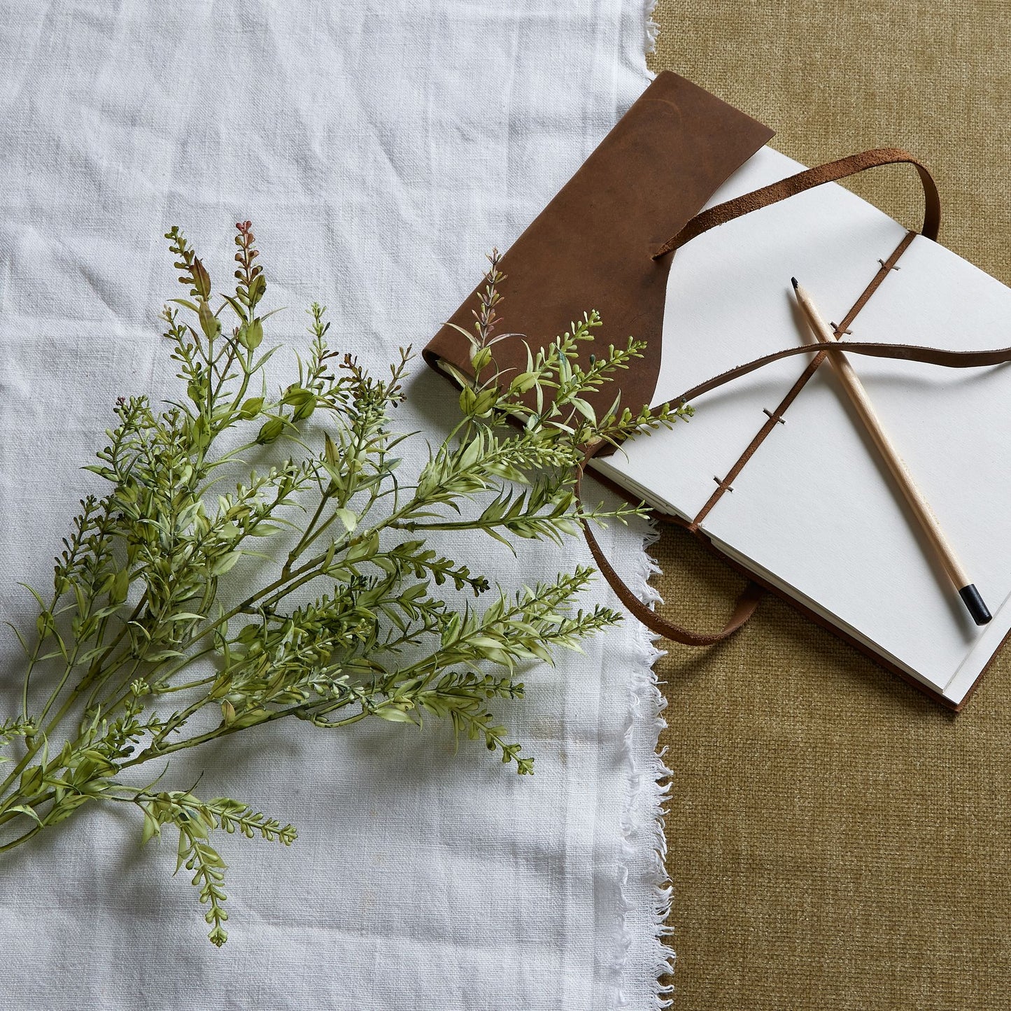 Broom stem in green with authentic organic texture, resting beside a leather-bound notebook and pencil on layered fabric backgrounds.