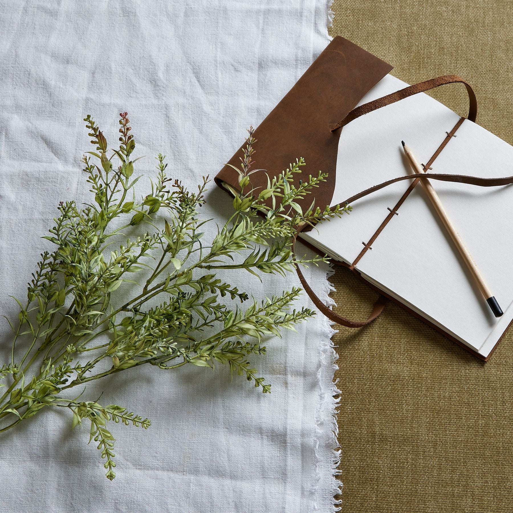 Broom stem in green with authentic organic texture, resting beside a leather-bound notebook and pencil on layered fabric backgrounds.