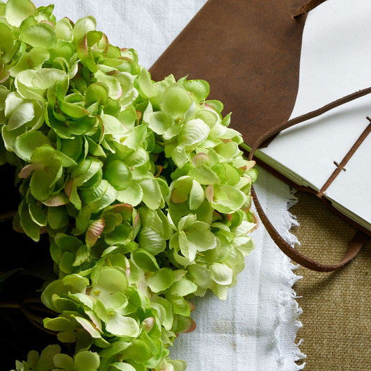 Lush green hydrangea bunch with hand-painted petals, arranged elegantly beside a notebook and rustic leather accents.