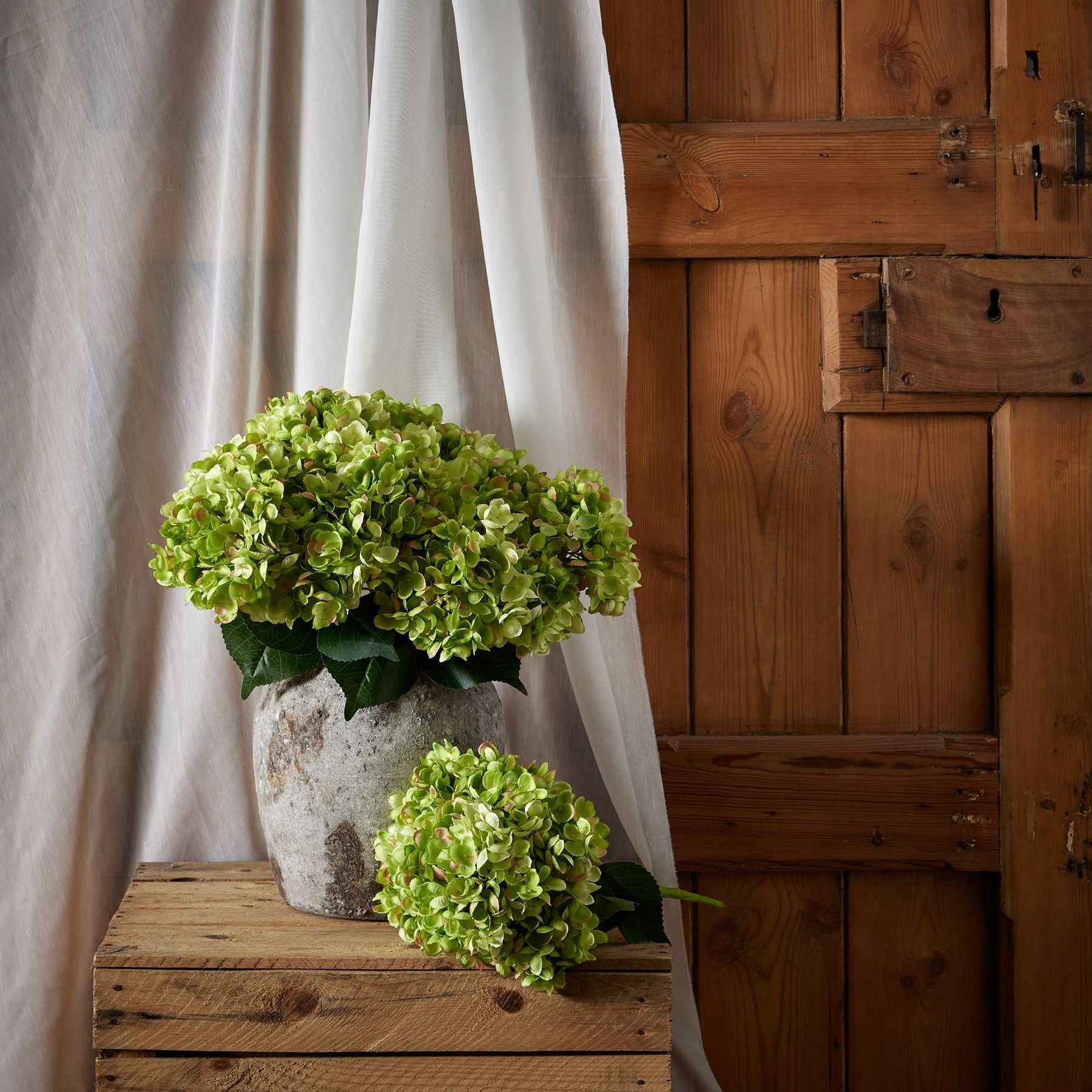 Hand-painted green hydrangea bunch in a textured gray vase, accented by rich leaves, set against warm wooden background.