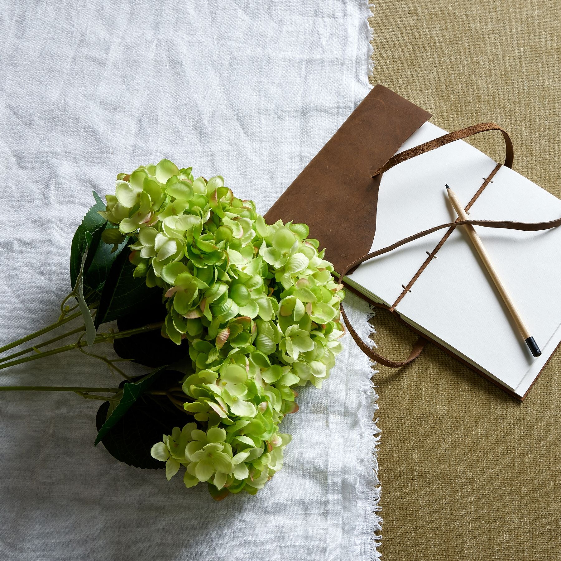 Artificial green hydrangea bunch with hand-painted petals, set against a white linen cloth and a rustic leather notebook.