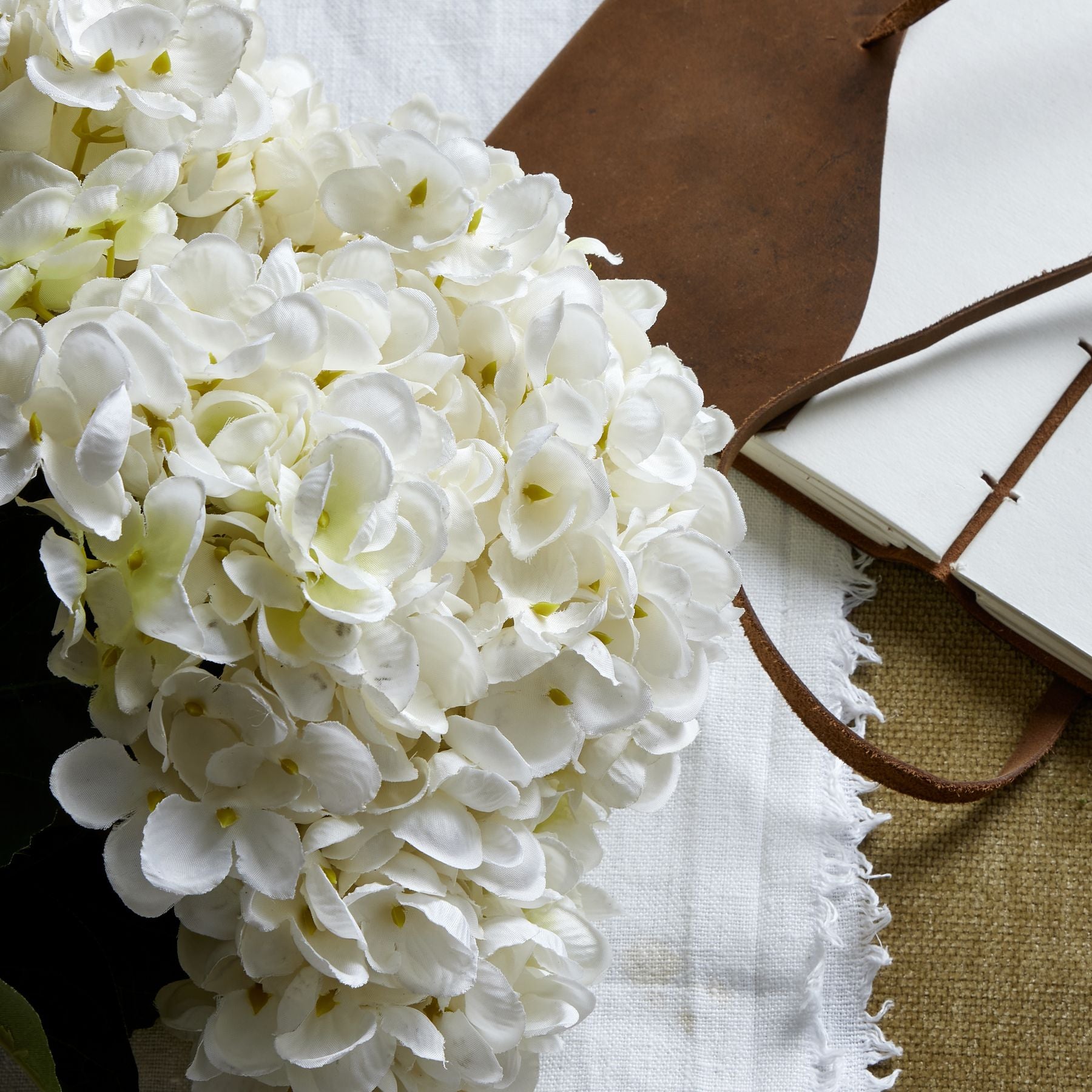 Pristine white hydrangea bunch with intricately crafted petals, laid on a textured background with a leather notebook and fabric.