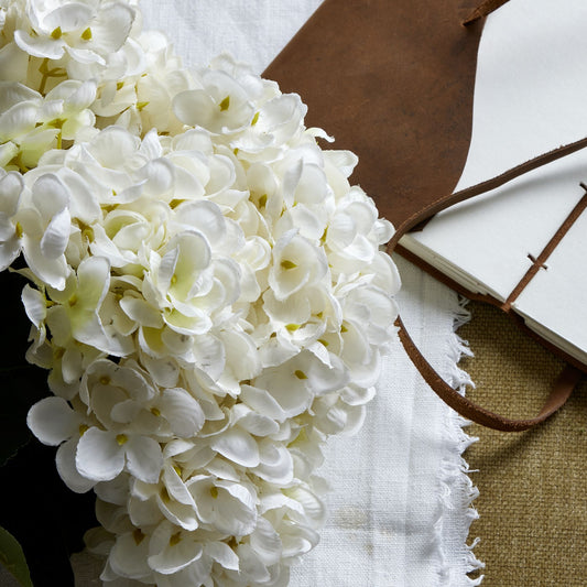 Pristine white hydrangea bunch with intricately crafted petals, laid on a textured background with a leather notebook and fabric.