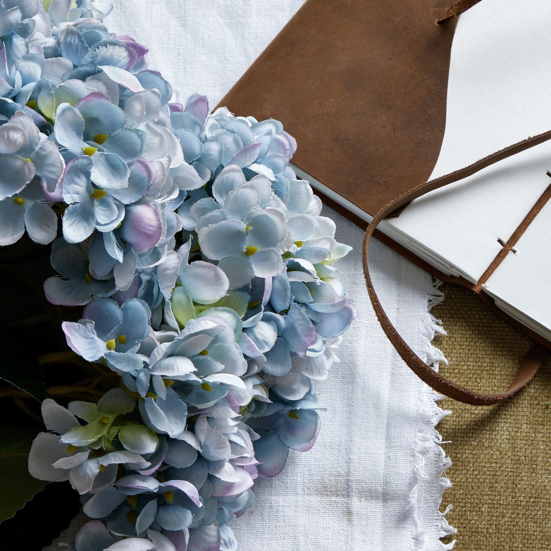 Lush blue hydrangea blooms with delicate details, paired with a brown leather notebook on textured fabric, evoking elegance.