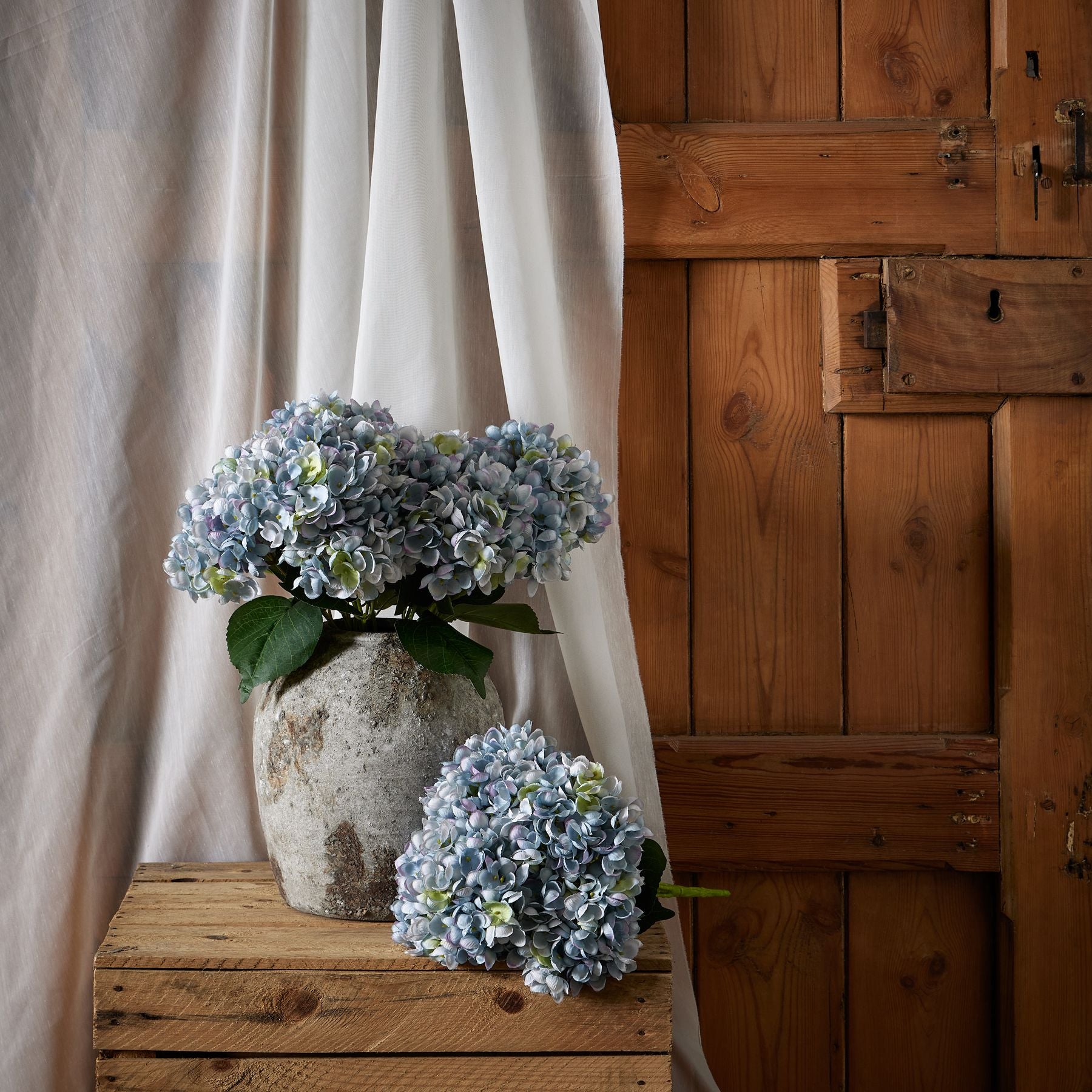 Lifelike blue hydrangea bunch in a textured vase, surrounded by soft white drapes and rustic wooden backdrop, creating an elegant ambiance.
