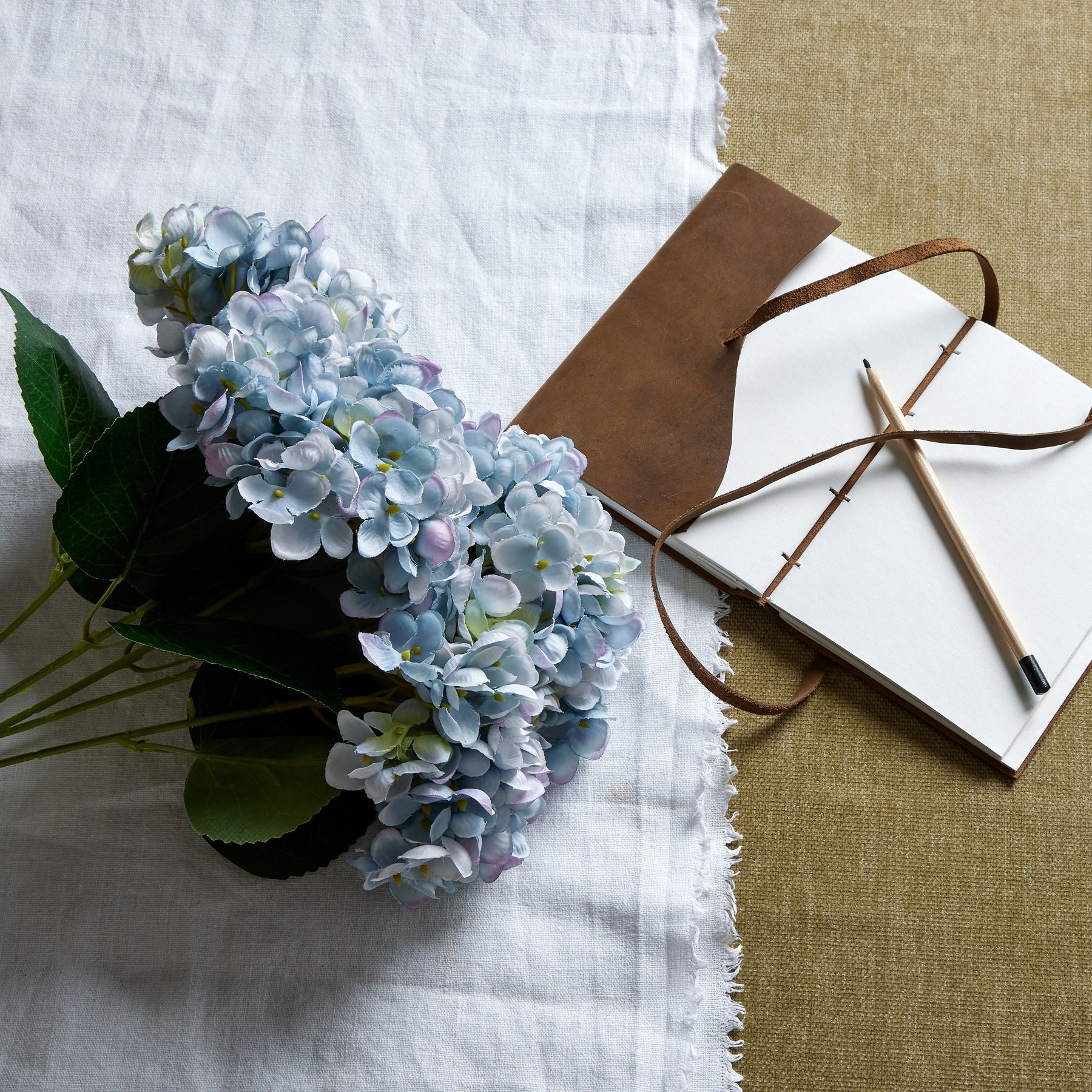 Lush blue hydrangea bunch with intricate blooms and green leaves, arranged alongside a brown leather notebook and pencil on a textured table.