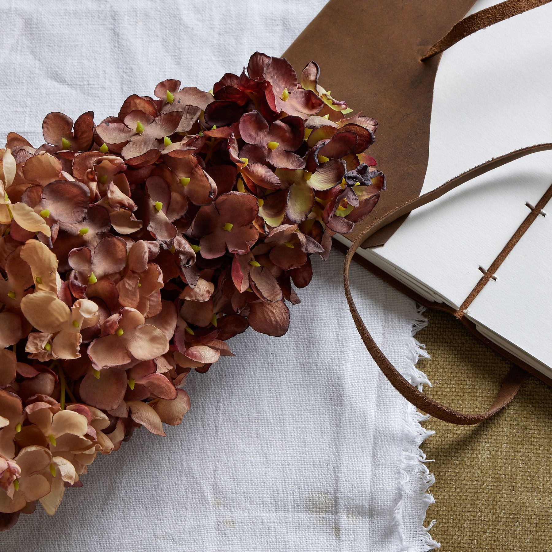 Deep burgundy hydrangea blooms with realistic detailing, arranged on a textured white cloth beside a leather-bound notebook.
