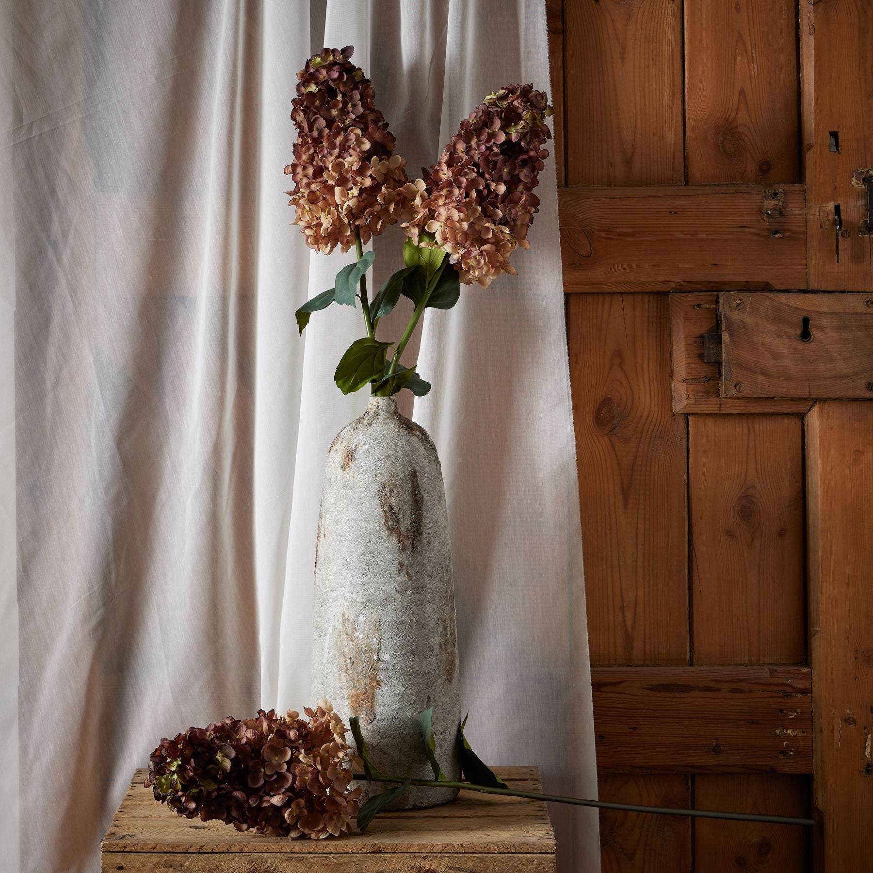 Burgundy Spear Hydrangea in a textured vase, featuring deep burgundy blooms and green leaves, set against a wooden backdrop.