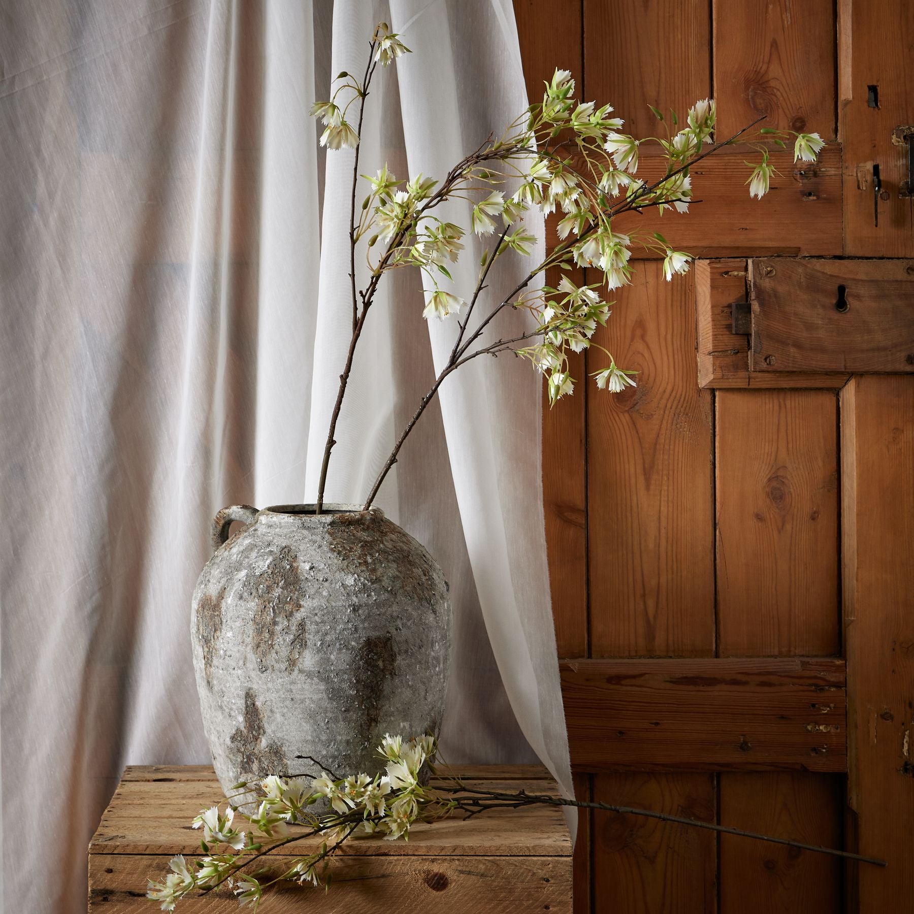 Elegant artificial Elaeocarpus stem in a rustic gray vase, featuring finely textured green leaves and delicate white flowers.