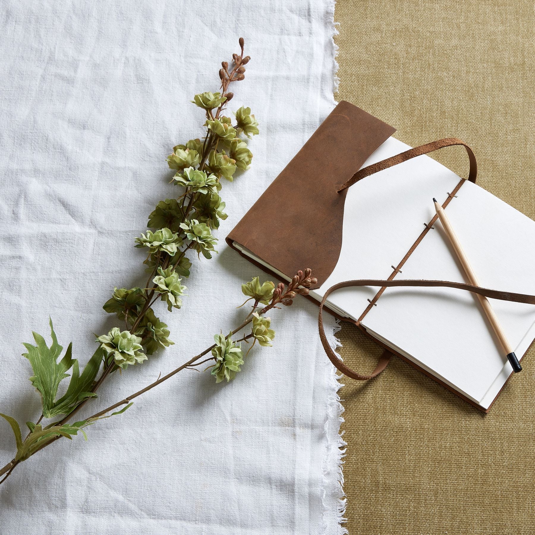 Slim green hops stem with delicate leaves, arranged alongside a leather-bound notebook and pencil on textured fabric backdrop.