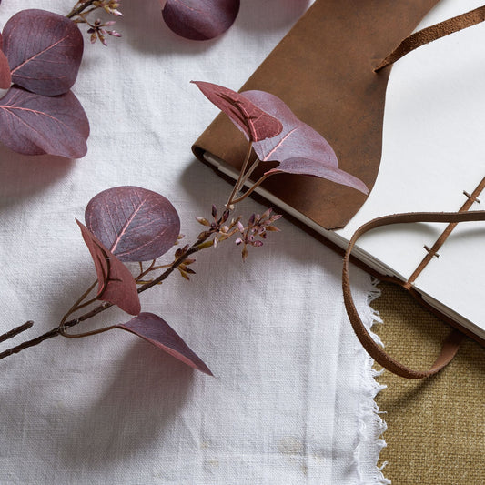 Burgundy berried eucalyptus stem with deep burgundy leaves and delicate berry accents, styled alongside a notebook on textured fabric.