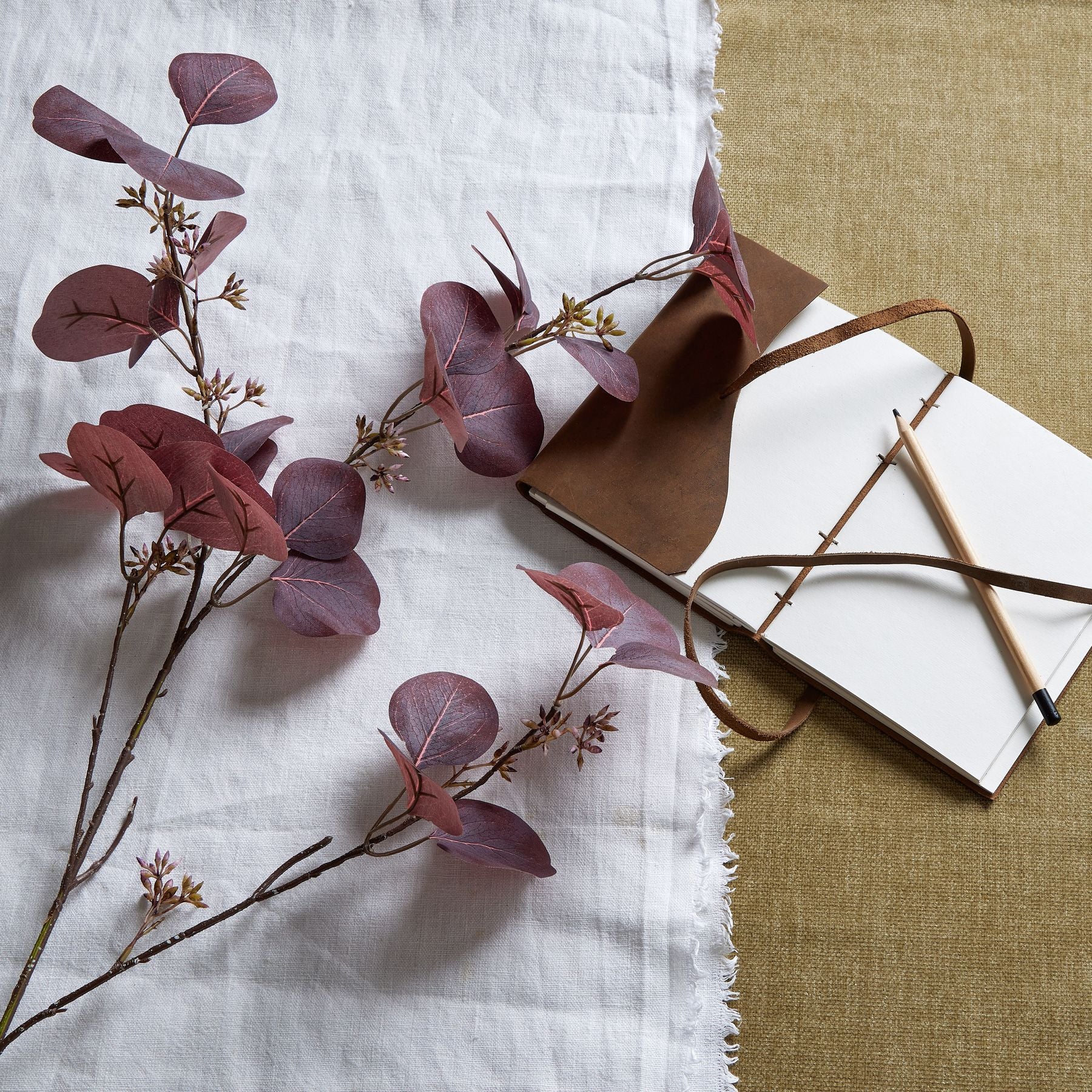 Burgundy berried eucalyptus stem features deep burgundy leaves and delicate berries, styled against textured fabric and a notebook.