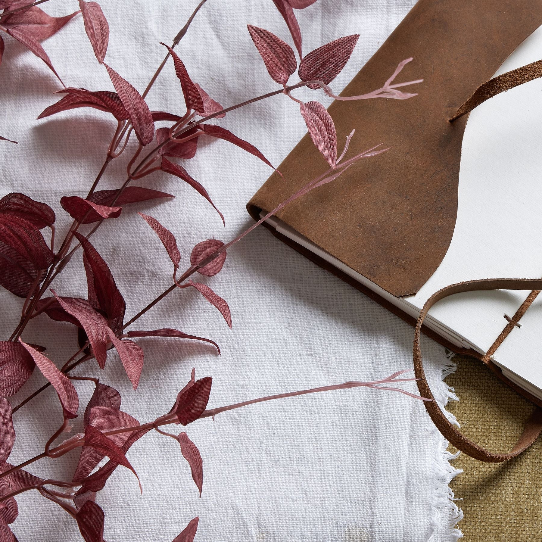 Rich burgundy leaves gracefully arranged beside a brown leather notebook on a textured white and linen background.