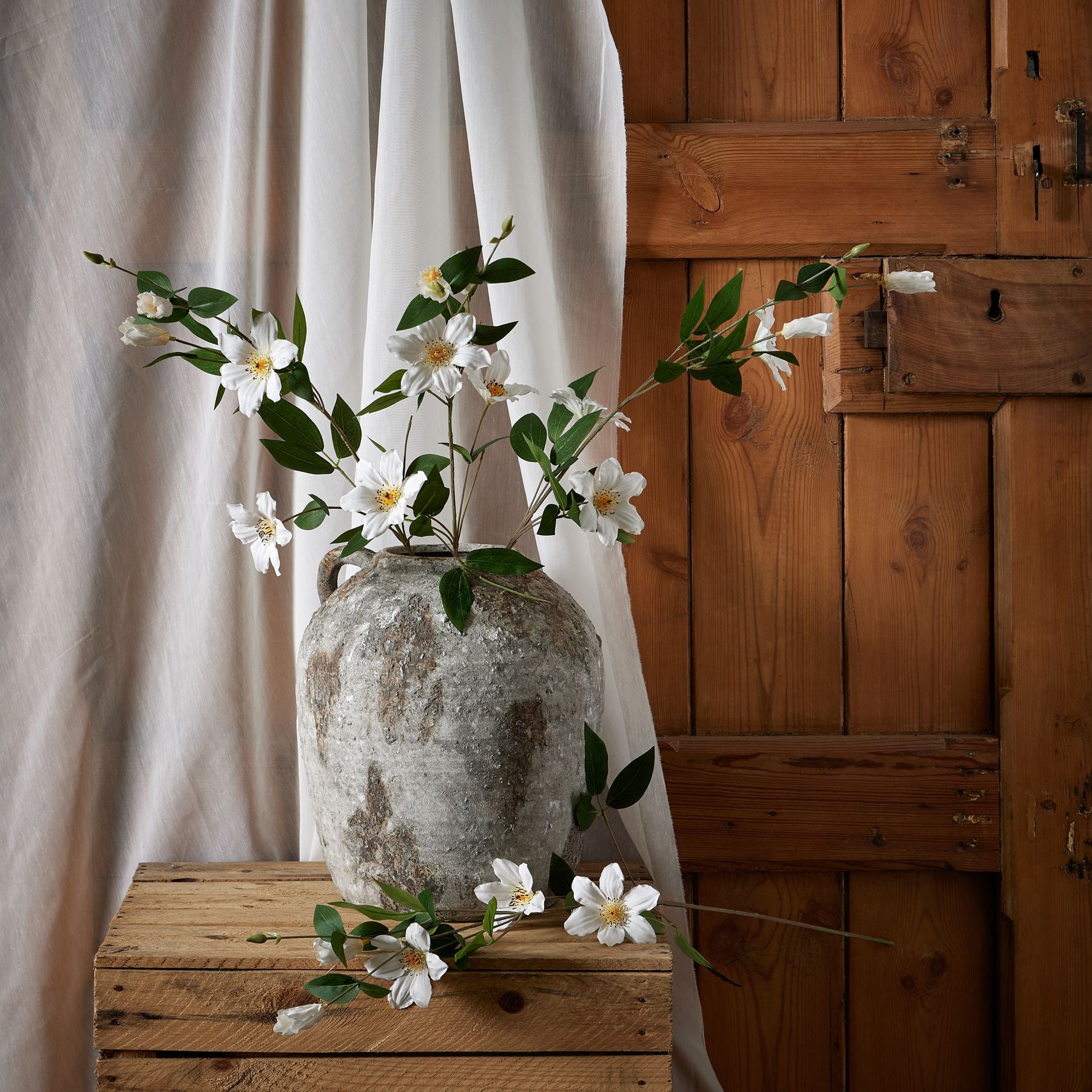 Elegant white clematis artificial flowers in a rustic gray vase, complemented by lush green leaves against a warm wooden backdrop.