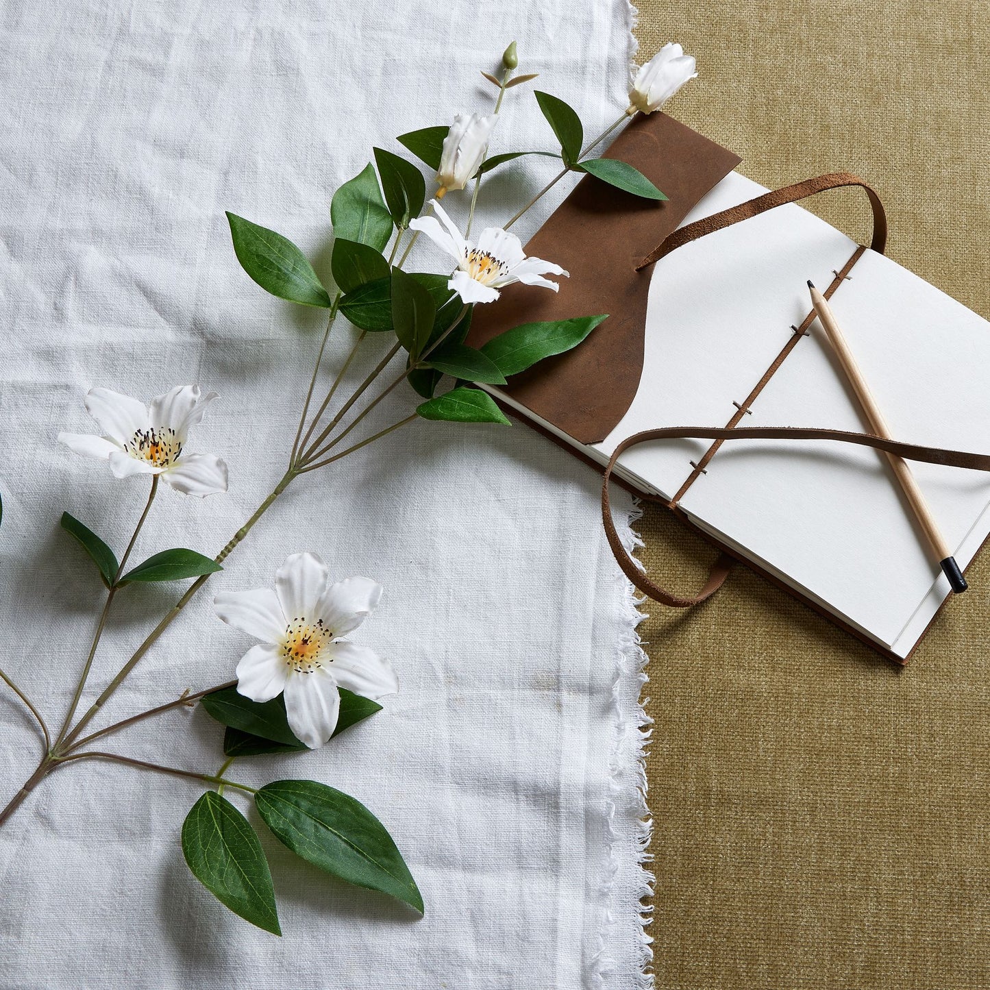 White Florida clematis stem with detailed blossoms and lush green leaves, arranged beside a notebook and pencil on textured fabric.
