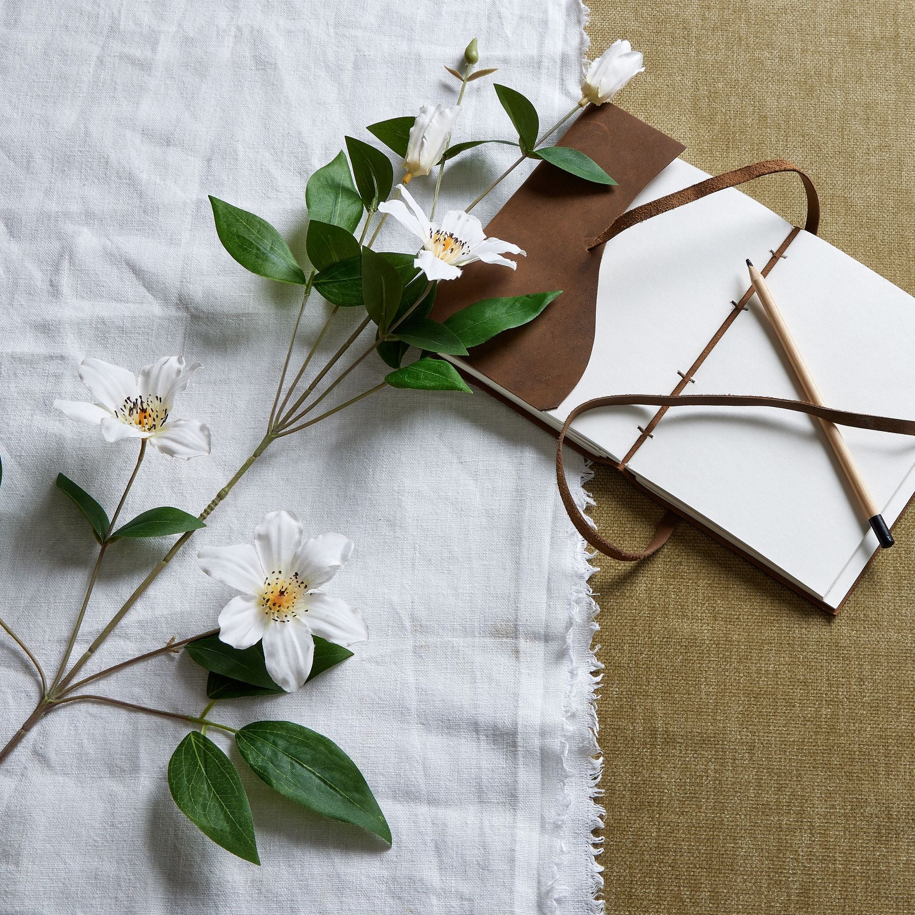 White Florida clematis stem with detailed blossoms and lush green leaves, arranged beside a notebook and pencil on textured fabric.