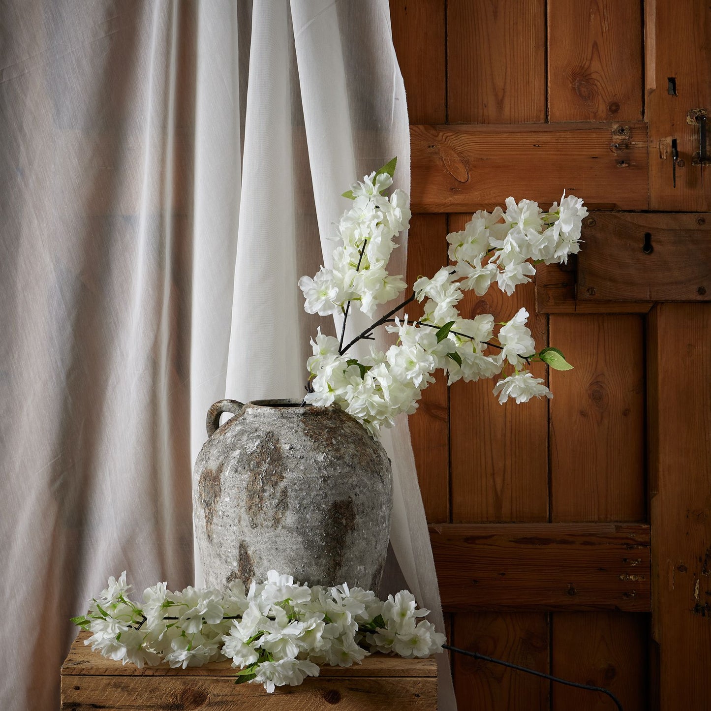 A large white cherry blossom stem with delicately crafted petals, arranged elegantly in a rustic gray vase against a wooden backdrop.
