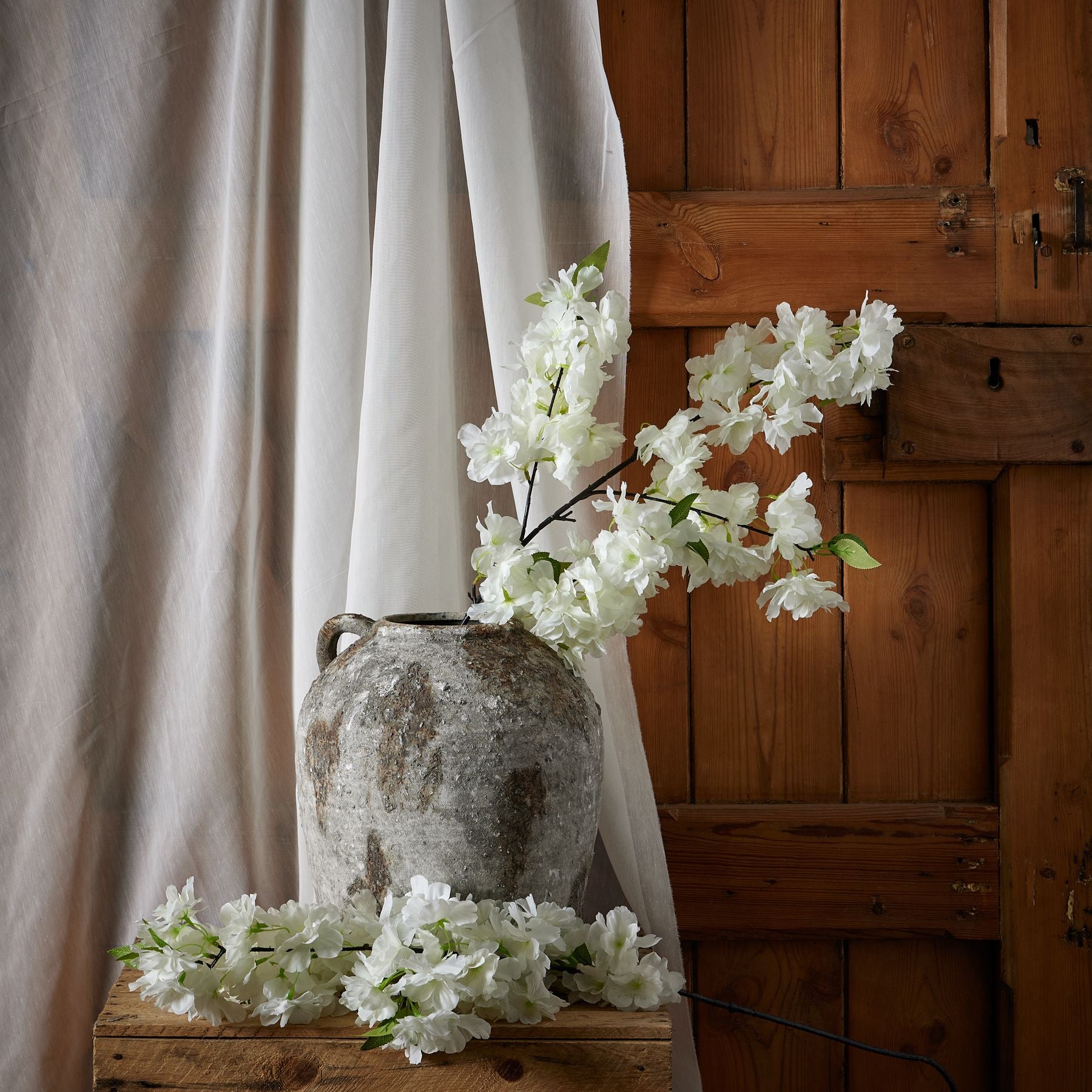 A large white cherry blossom stem with delicately crafted petals, arranged elegantly in a rustic gray vase against a wooden backdrop.