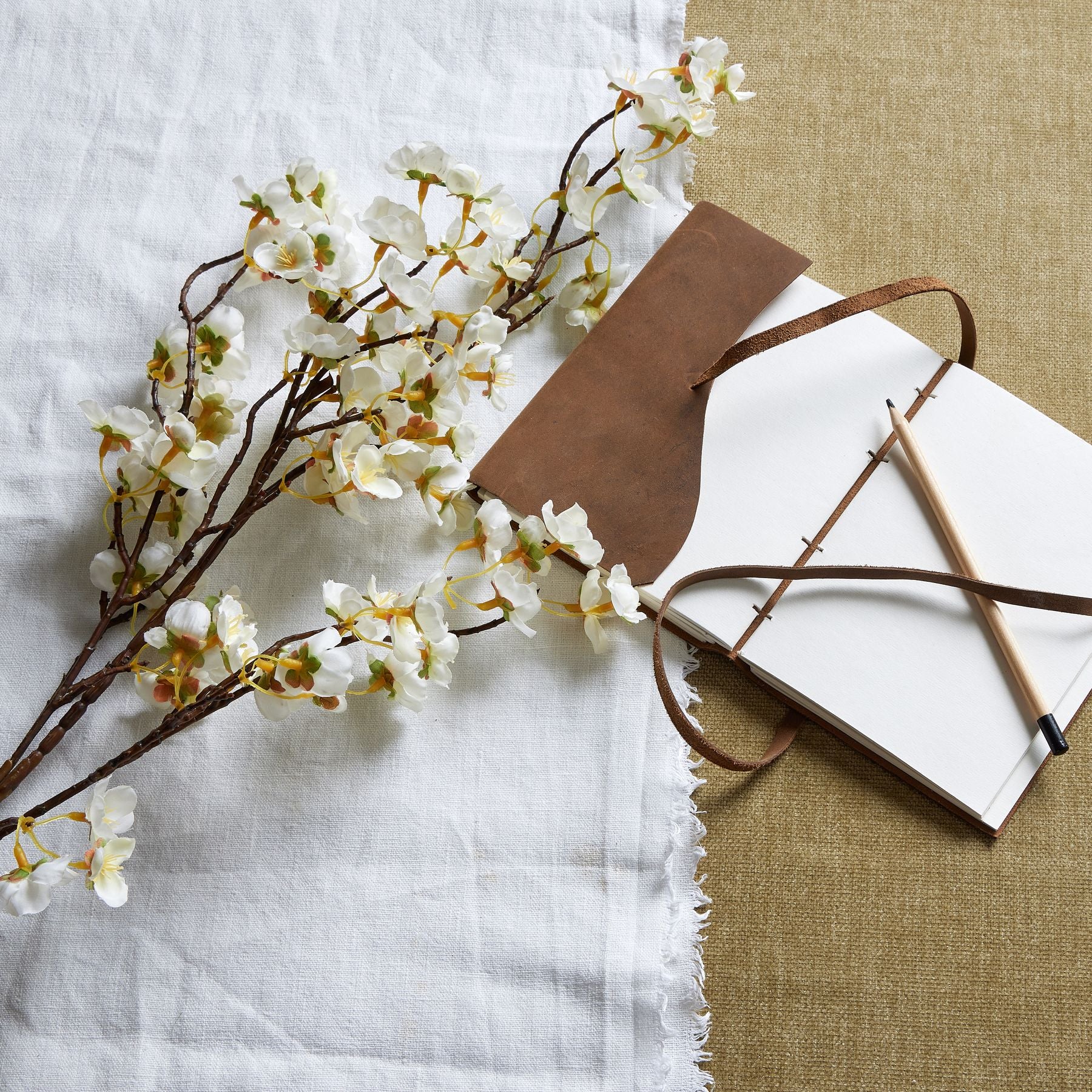 Delicate white Japanese blossoms on elegant branches, styled with a leather-bound notebook and pencil on textured fabrics.