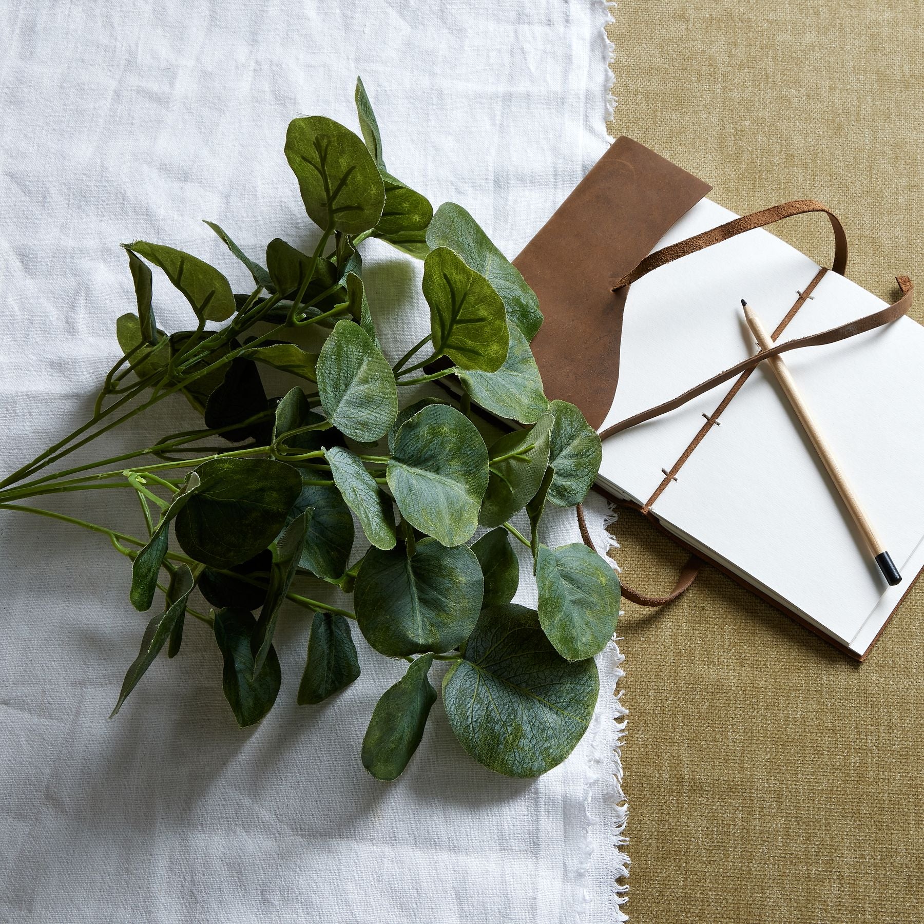 Lifelike short eucalyptus bunch with rich green leaves, arranged on a textured linen backdrop beside a notebook and pencil.