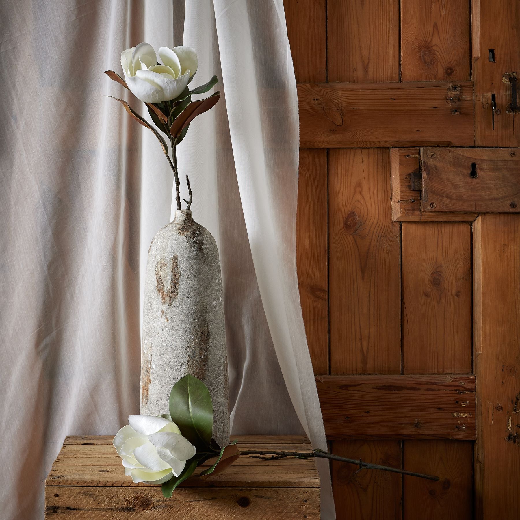 Ethereal white magnolia stem with detailed petals rests in a rustic vase against a backdrop of soft fabric and wooden panels.