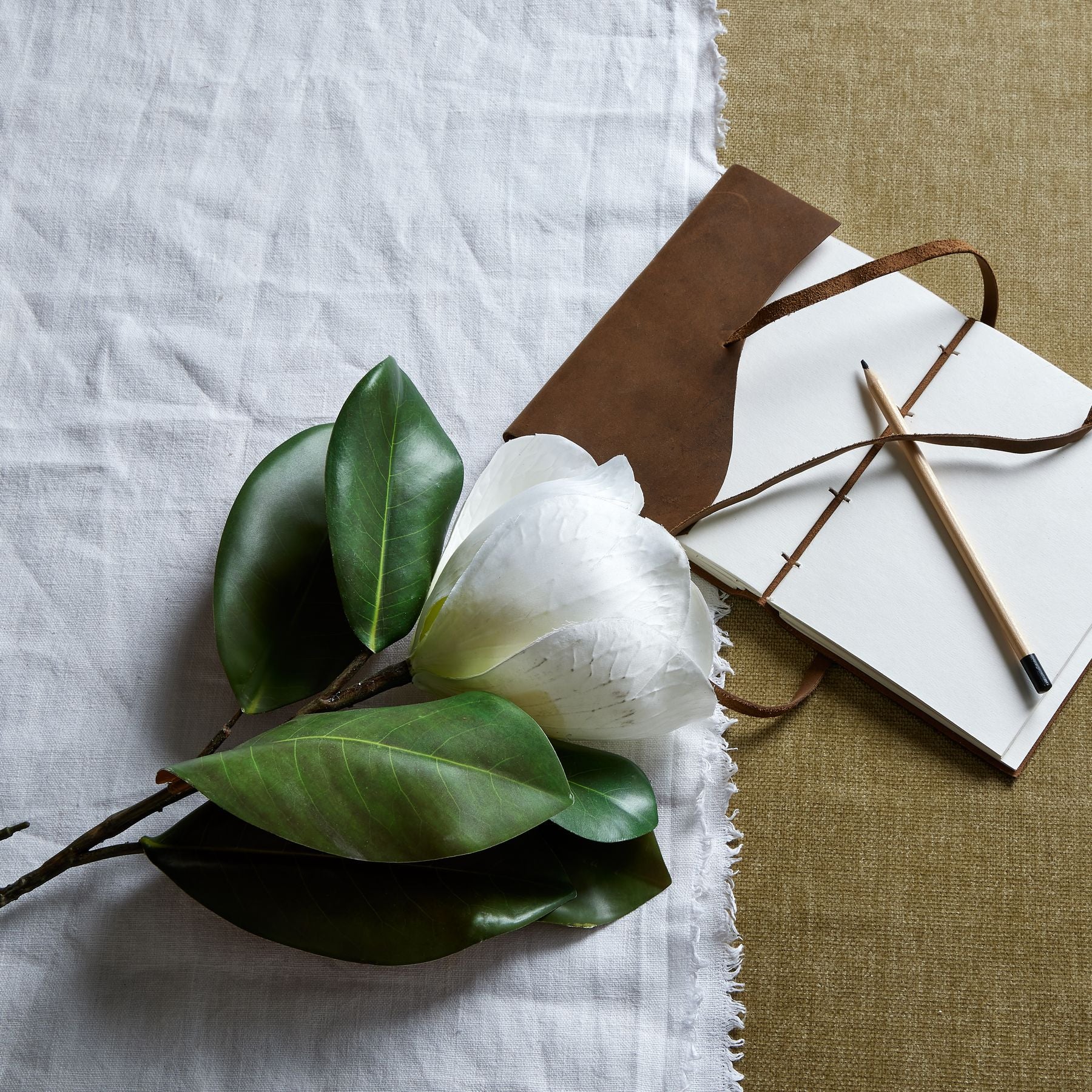 Elegant white magnolia stem with detailed petals and green leaves, set against white linen and a textured background.