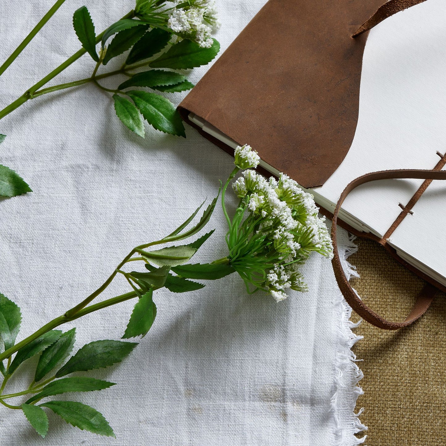 Detailed artificial white cow parsley stem with lace-like blooms, green leaves, and a rustic notebook on a white fabric background.