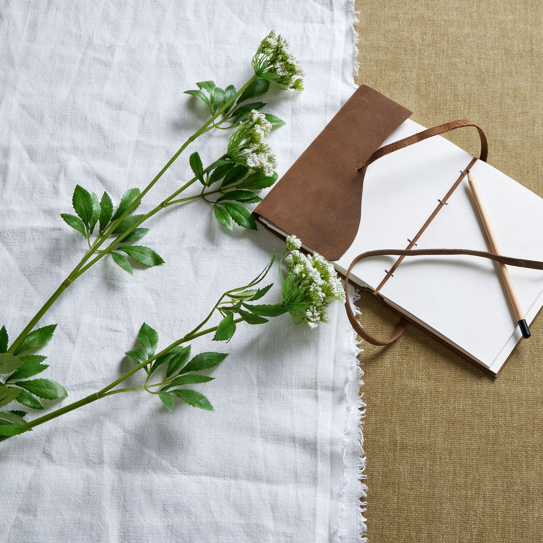 Delicate white blooms of faux cow parsley stem alongside green foliage, resting on a textured white and beige background with a brown notebook.