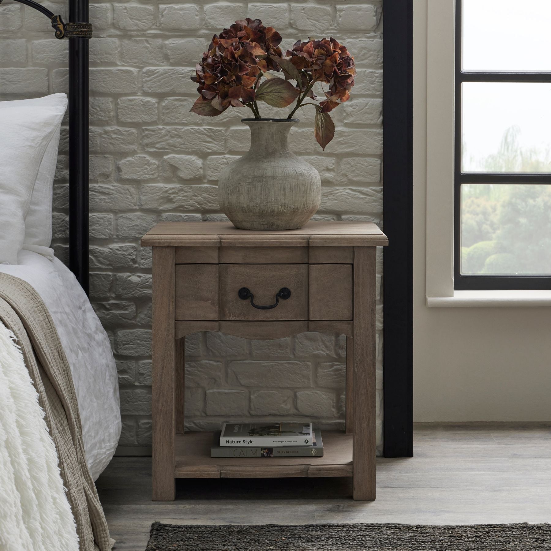 Rustic wooden side table with a washed finish, featuring an elegant drawer, shelf, and black iron handles, adorned with a vase of dried flowers.