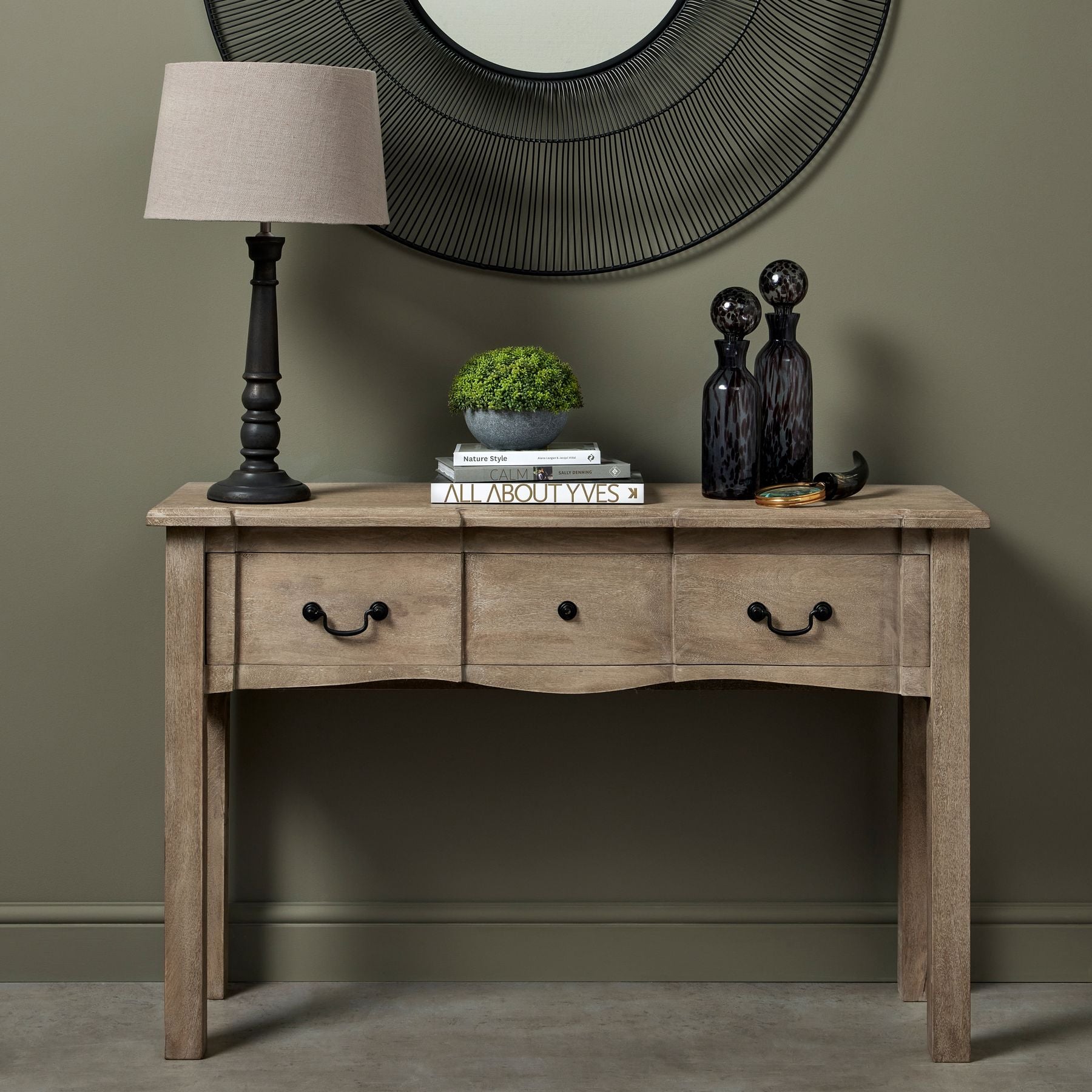 French provincial console table with a washed wood finish, black iron handles, and decorative accessories on top.