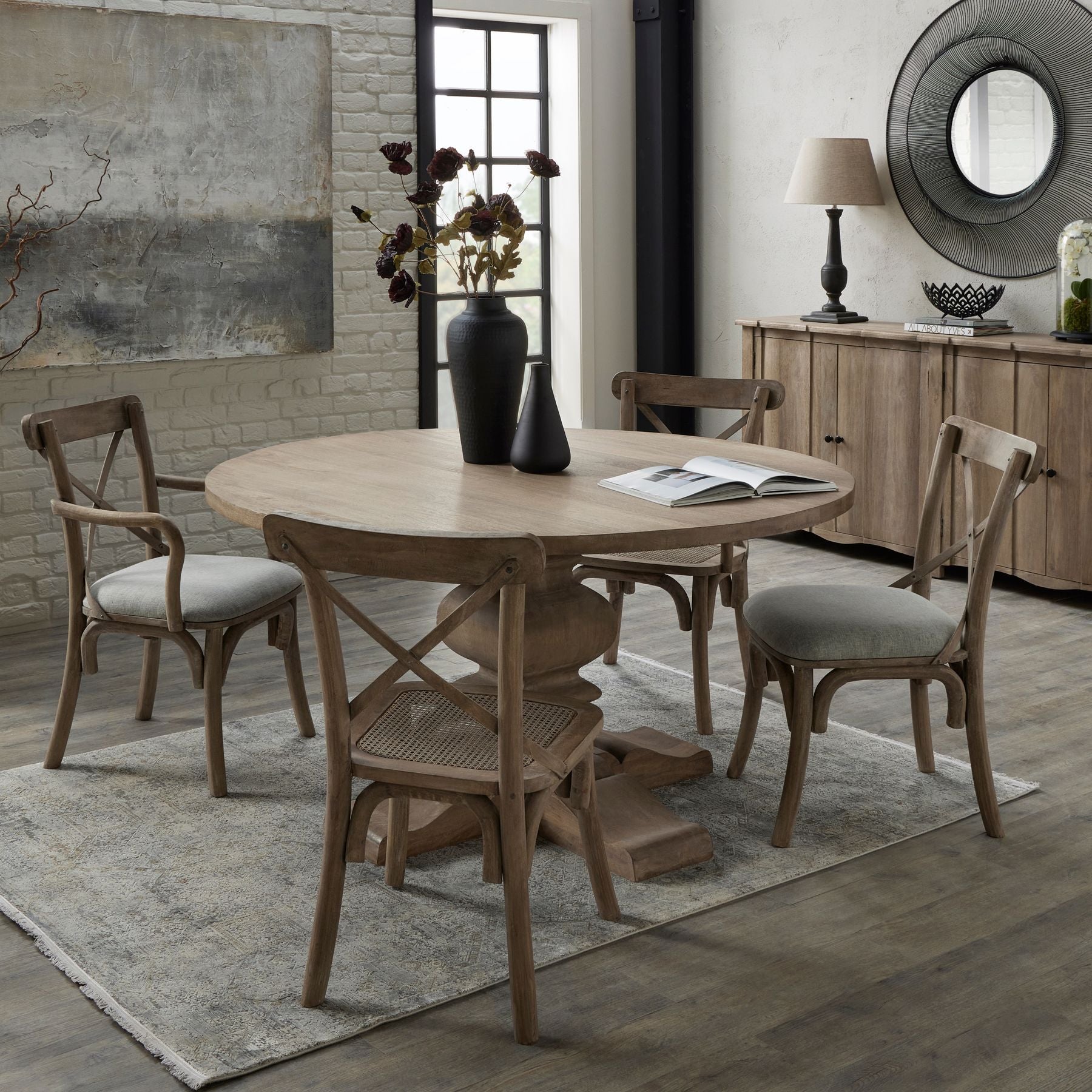 Round wooden pedestal dining table with a washed finish, complemented by light grey upholstered chairs, set in an elegant dining space.