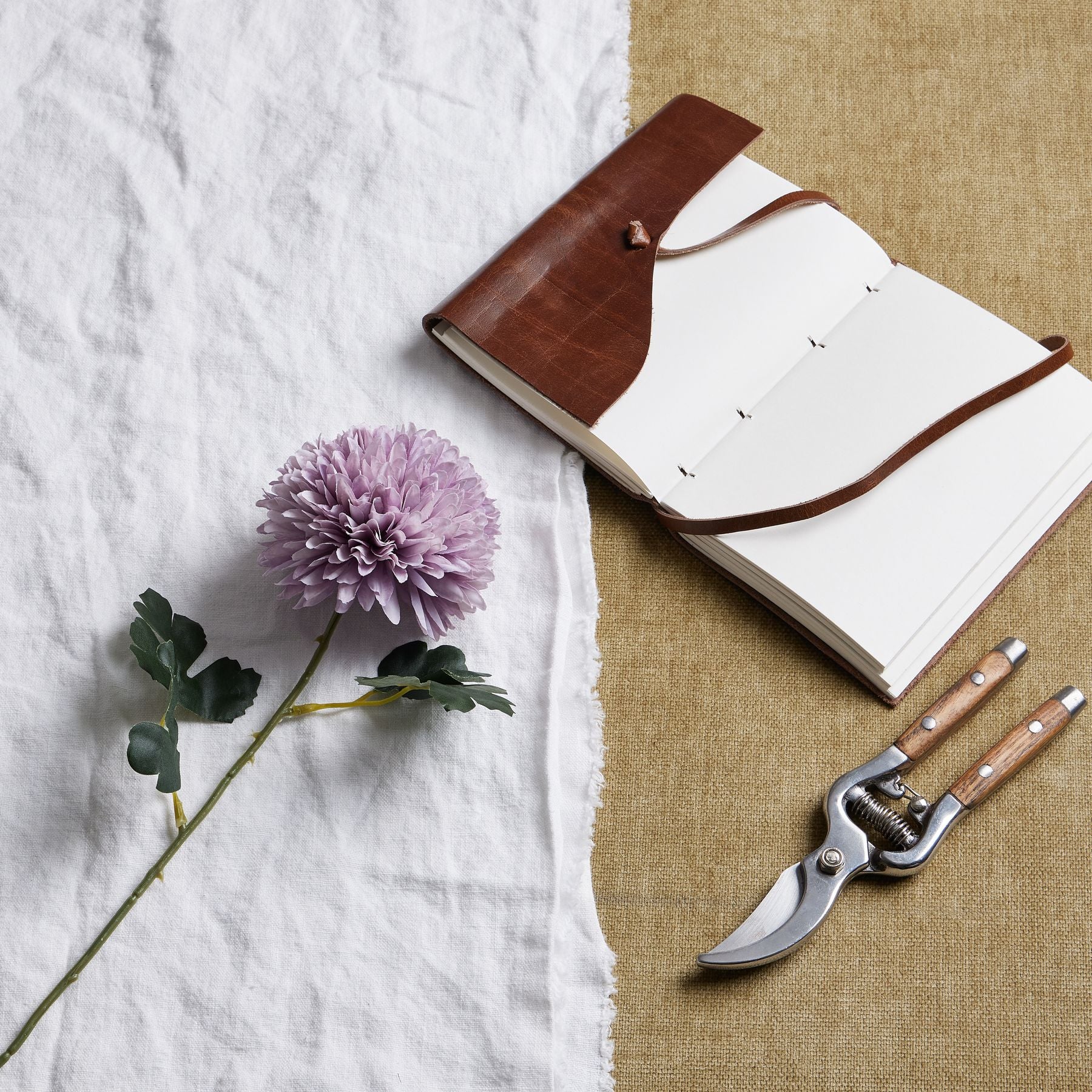 Light purple chrysanthemum with layered petals beside a leather notebook and garden shears on a textured surface.