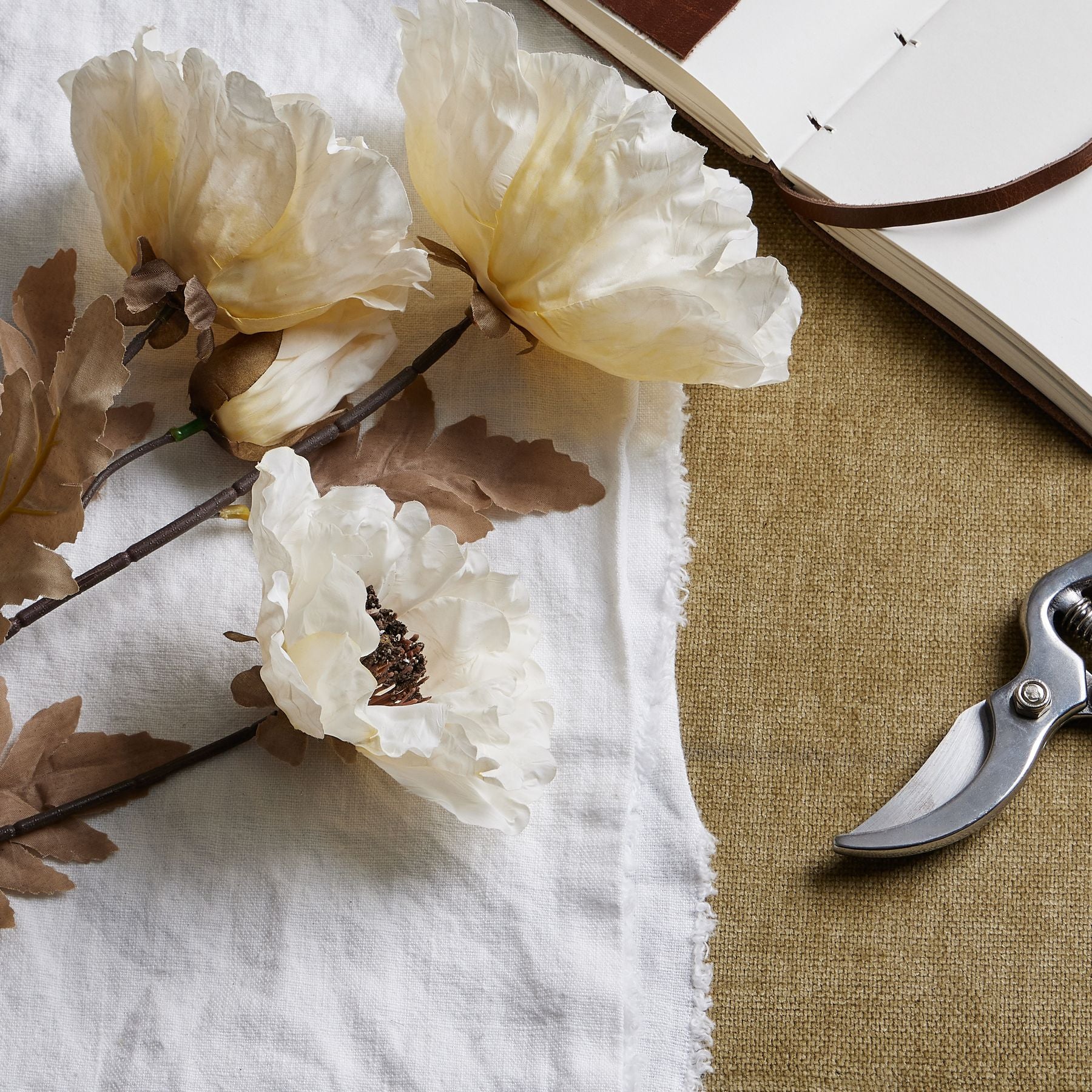 Large off-white poppy stem with subtle coffee accents, resting on a white cloth, alongside a notebook and gardening shears.