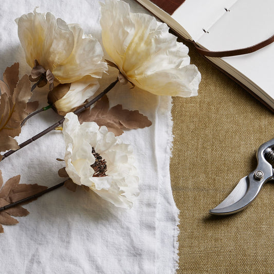 Large off-white poppy stem with subtle coffee accents, resting on a white cloth, alongside a notebook and gardening shears.
