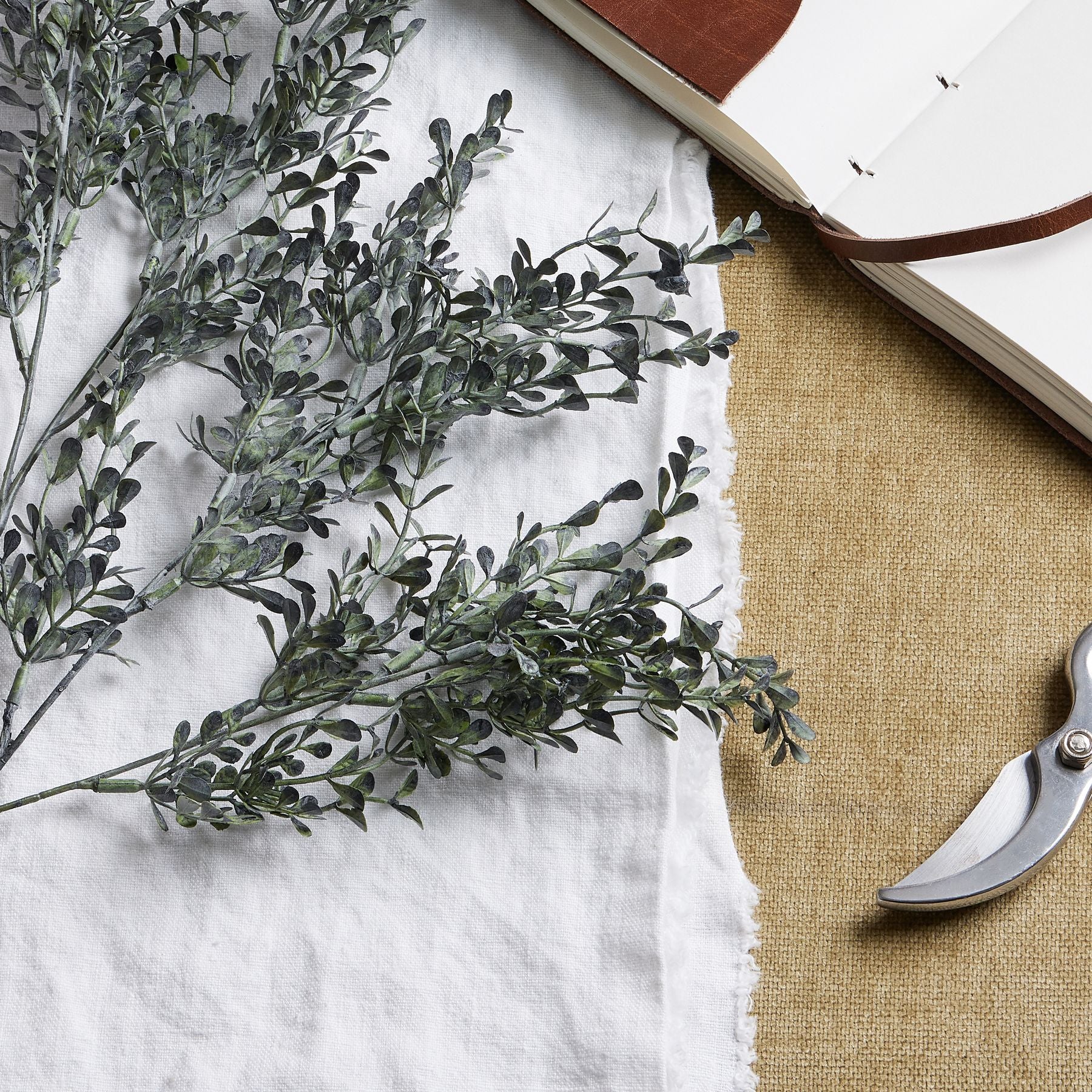 Elegant blackened grass spray stem featuring realistic green and black foliage, resting on a textured beige surface near a notebook and scissors.
