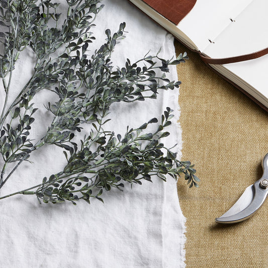 Elegant blackened grass spray stem featuring realistic green and black foliage, resting on a textured beige surface near a notebook and scissors.