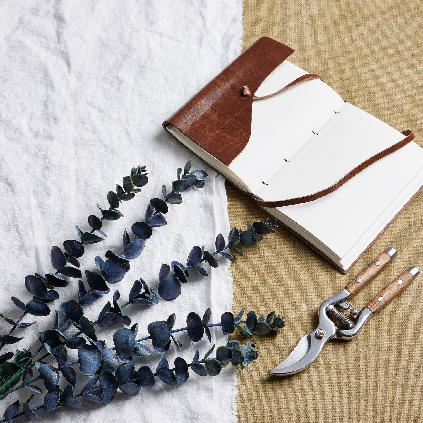 Large blue eucalyptus stem with rich tones, showcasing realistic leaves, next to a leather notebook and garden shears on textured fabric.