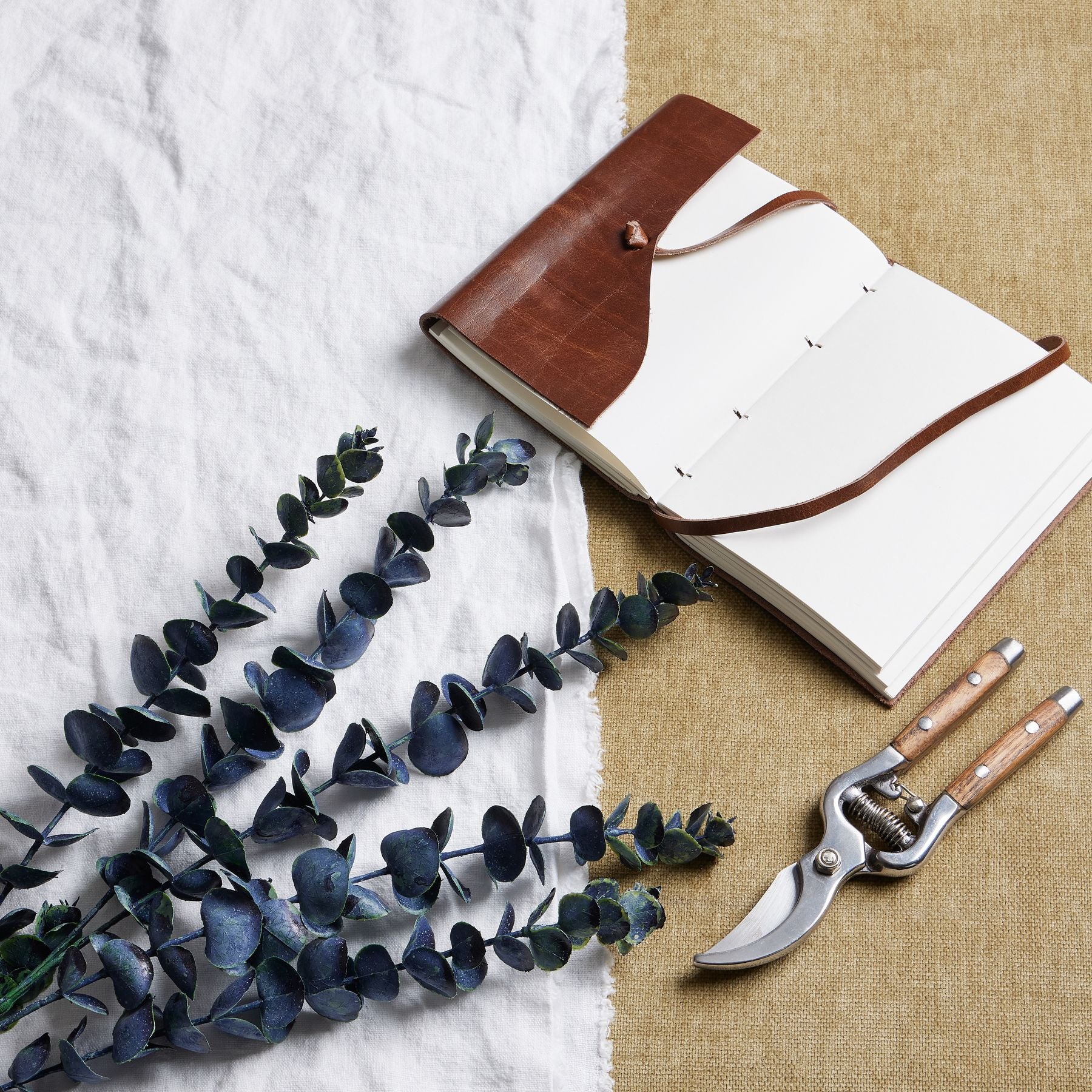 Large blue eucalyptus stem with rich tones, showcasing realistic leaves, next to a leather notebook and garden shears on textured fabric.