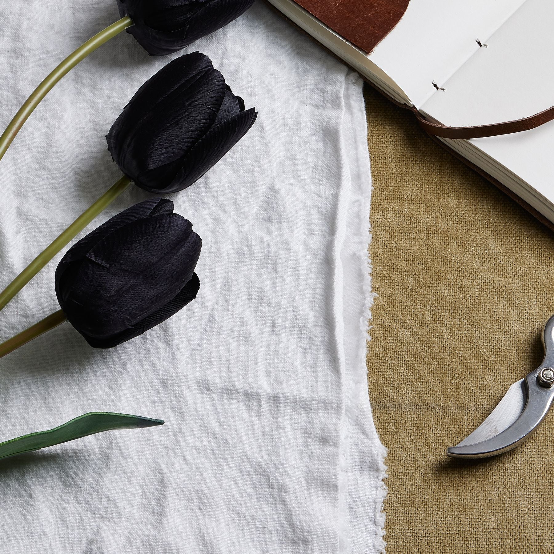 Tall black artificial tulip stems with sleek petals and green leaves, arranged on white fabric beside a notebook and scissors.